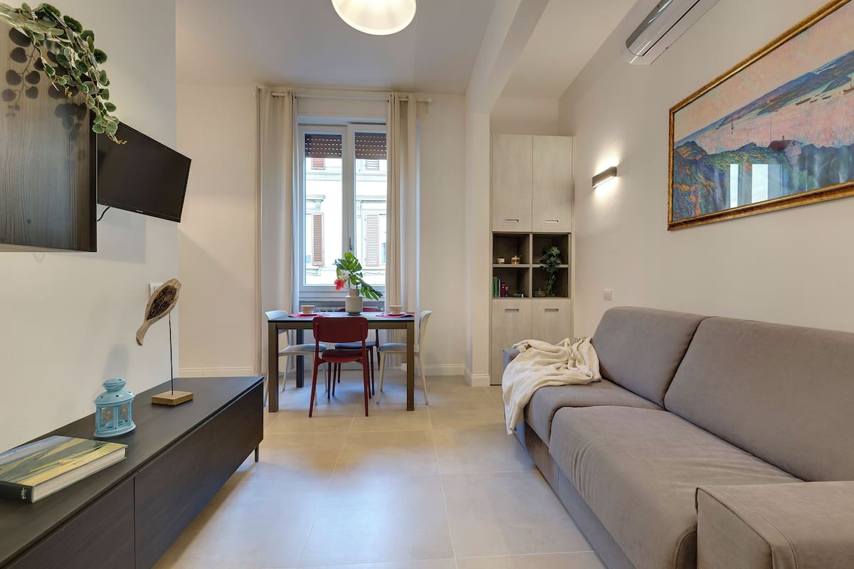 A modern living room with a sofa, a dining area with a table and chairs, a TV mounted on the wall, and a painting above the sofa.