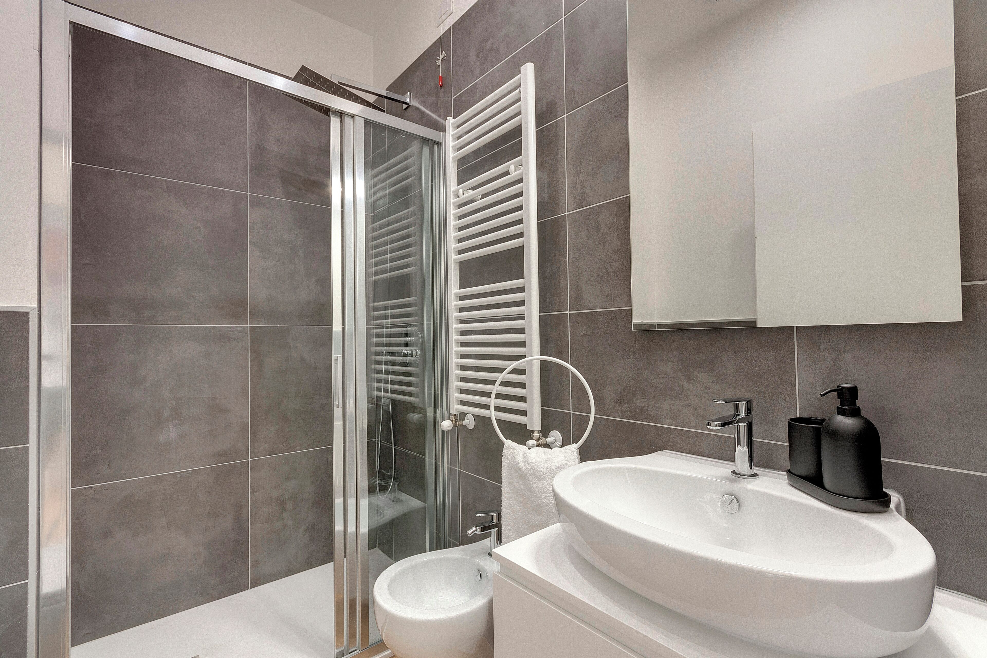 Un baño moderno con cabina de ducha de vidrio, un lavabo blanco con dispensador de jabón negro y un perchero para toallas.