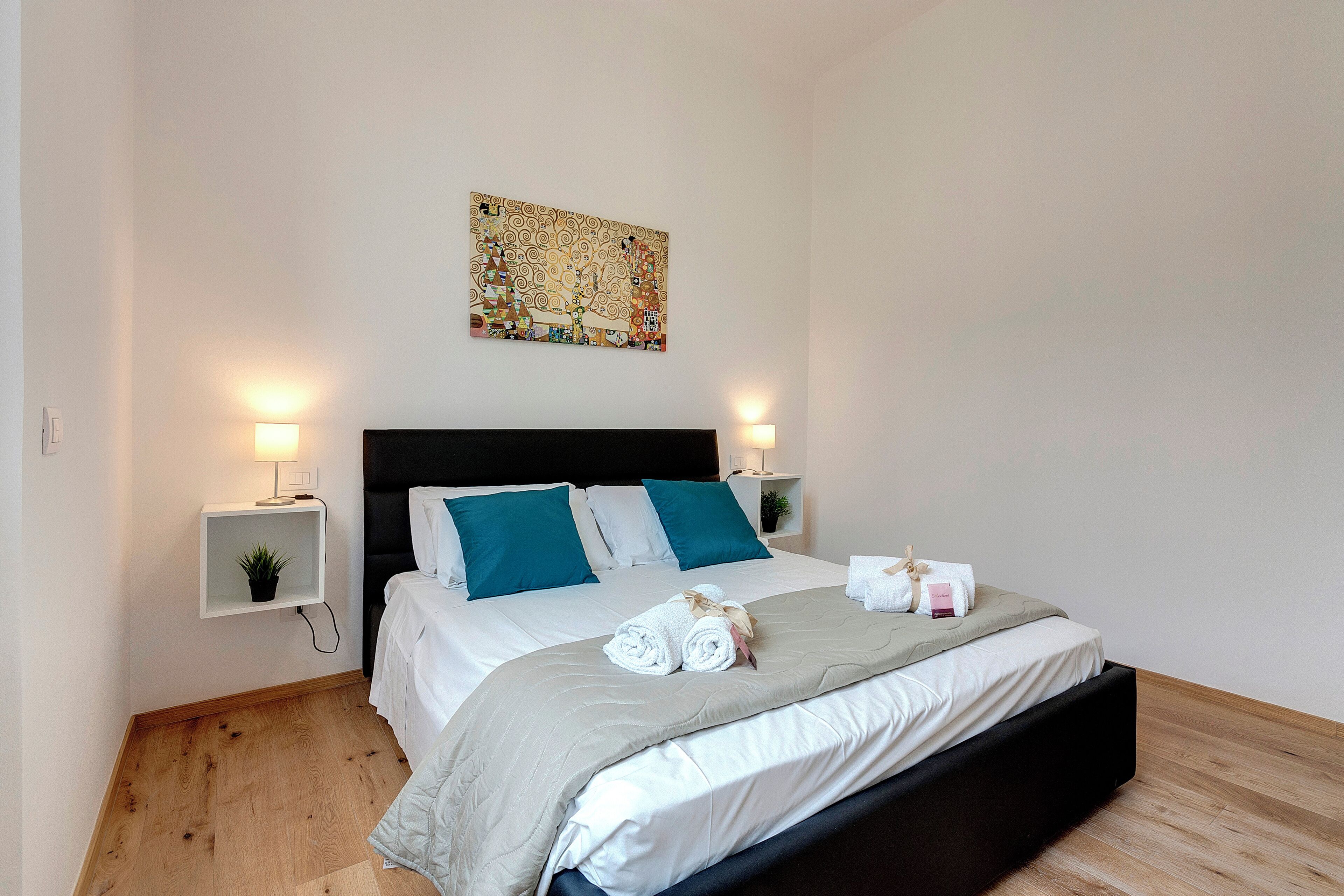 Cama bem arrumada com cabeceira preta, duas almofadas azul-turquesa e um cobertor branco. Um criado-mudo pequeno com uma luminária e uma planta. Um quadro está pendurado na parede acima da cama.
