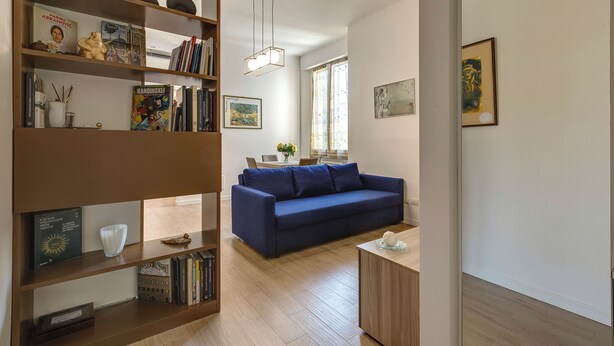A living room with a blue sofa, wooden shelves with books and decorative items, a small table with a vase, and a dining area in the background. - Mamo Florence - Cavour Apartments (Florence)