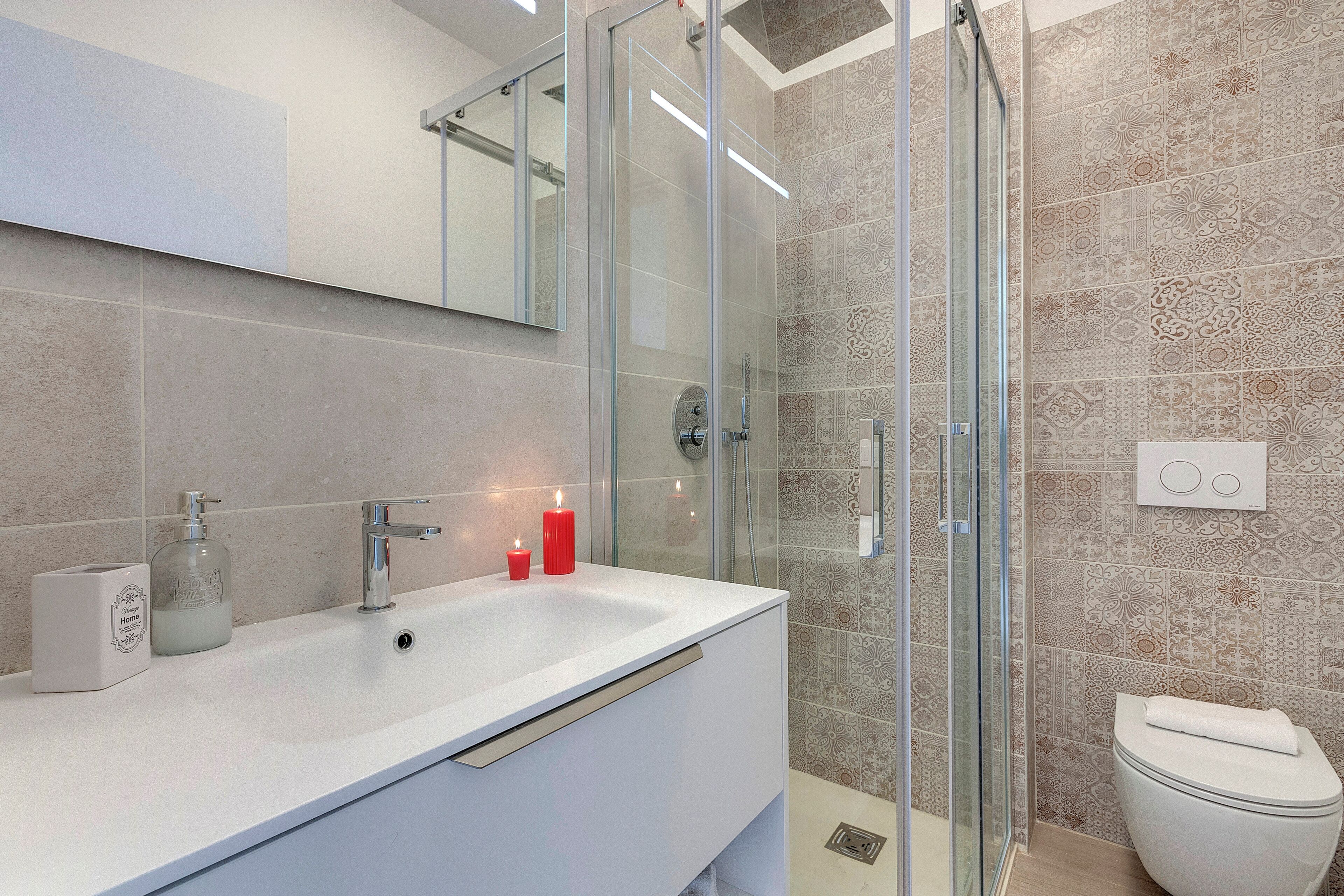 Un baño moderno con ducha envuelta en vidrio, un lavabo blanco con espejo arriba y baldosas estampadas en las paredes.