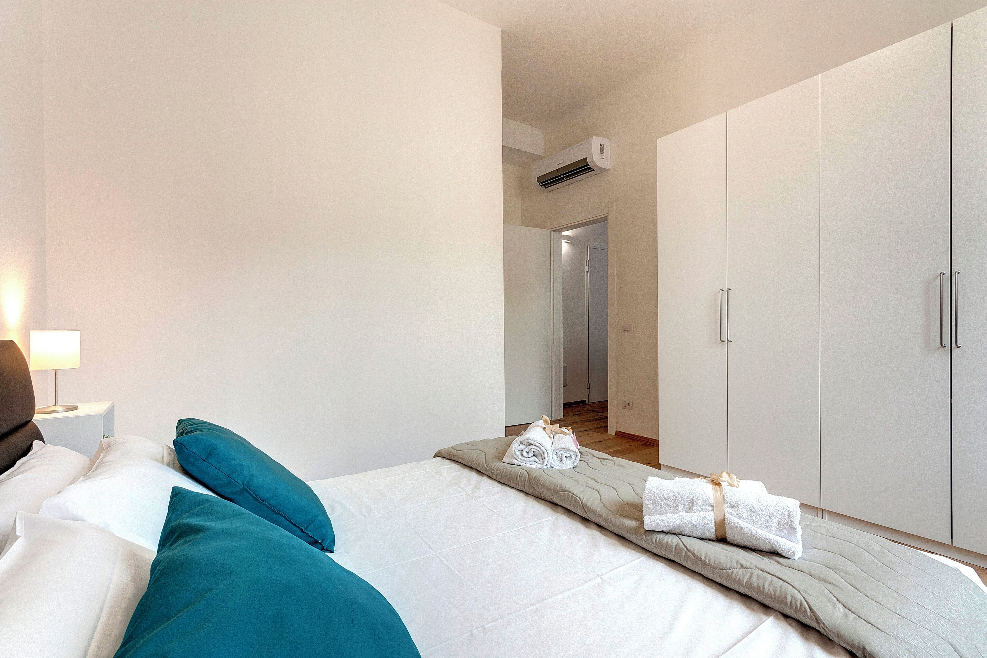 A hotel room with a bed, pillows, a nightstand, a lamp, and a wardrobe.