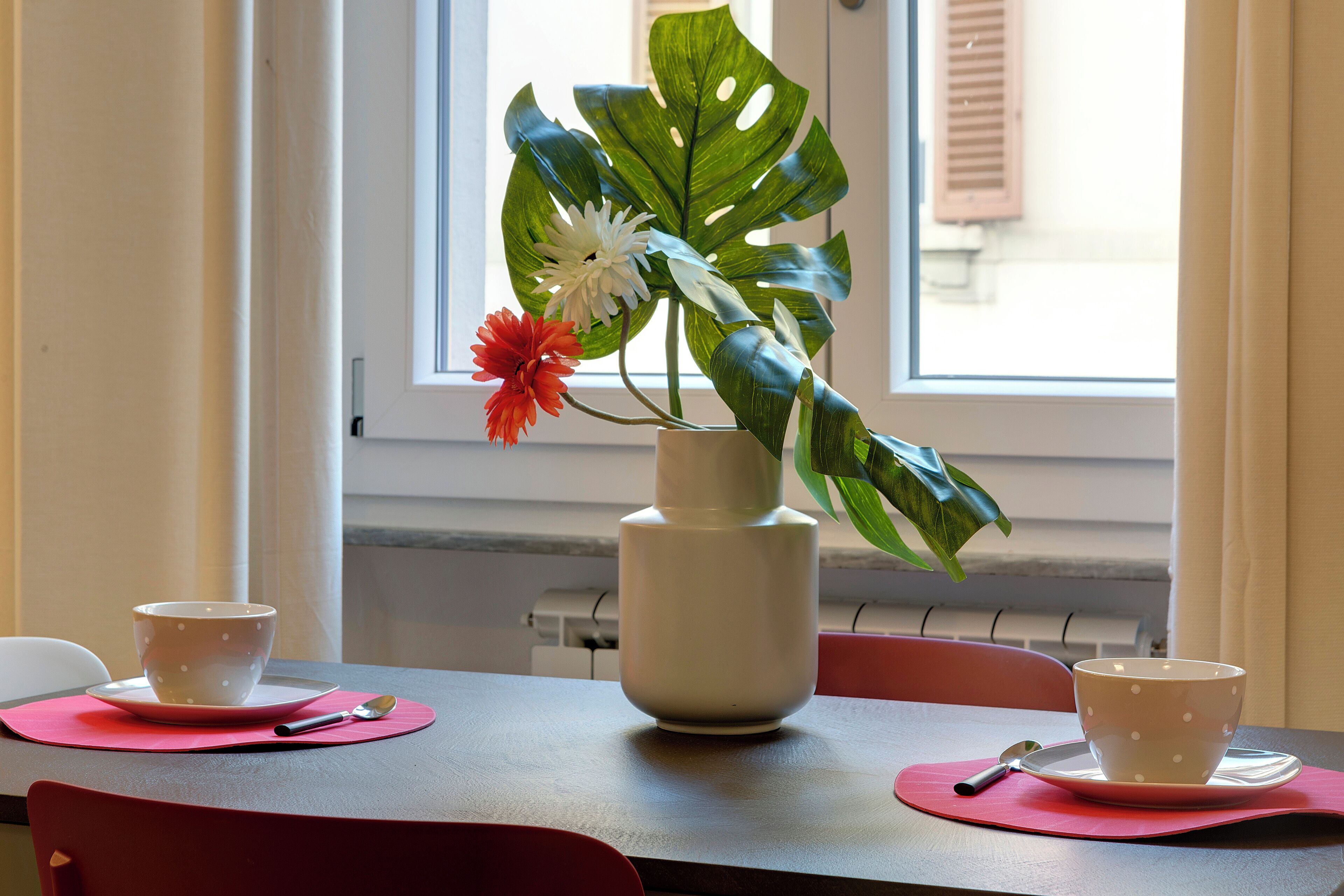 Una mesa con dos tazas blancas sobre individuales rosa, un jarrón blanco con hojas y flores verdes, y una ventana con persianas.