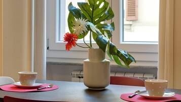 Una mesa con dos tazas blancas sobre individuales rosa, un jarrón blanco con hojas y flores verdes, y una ventana con persianas.