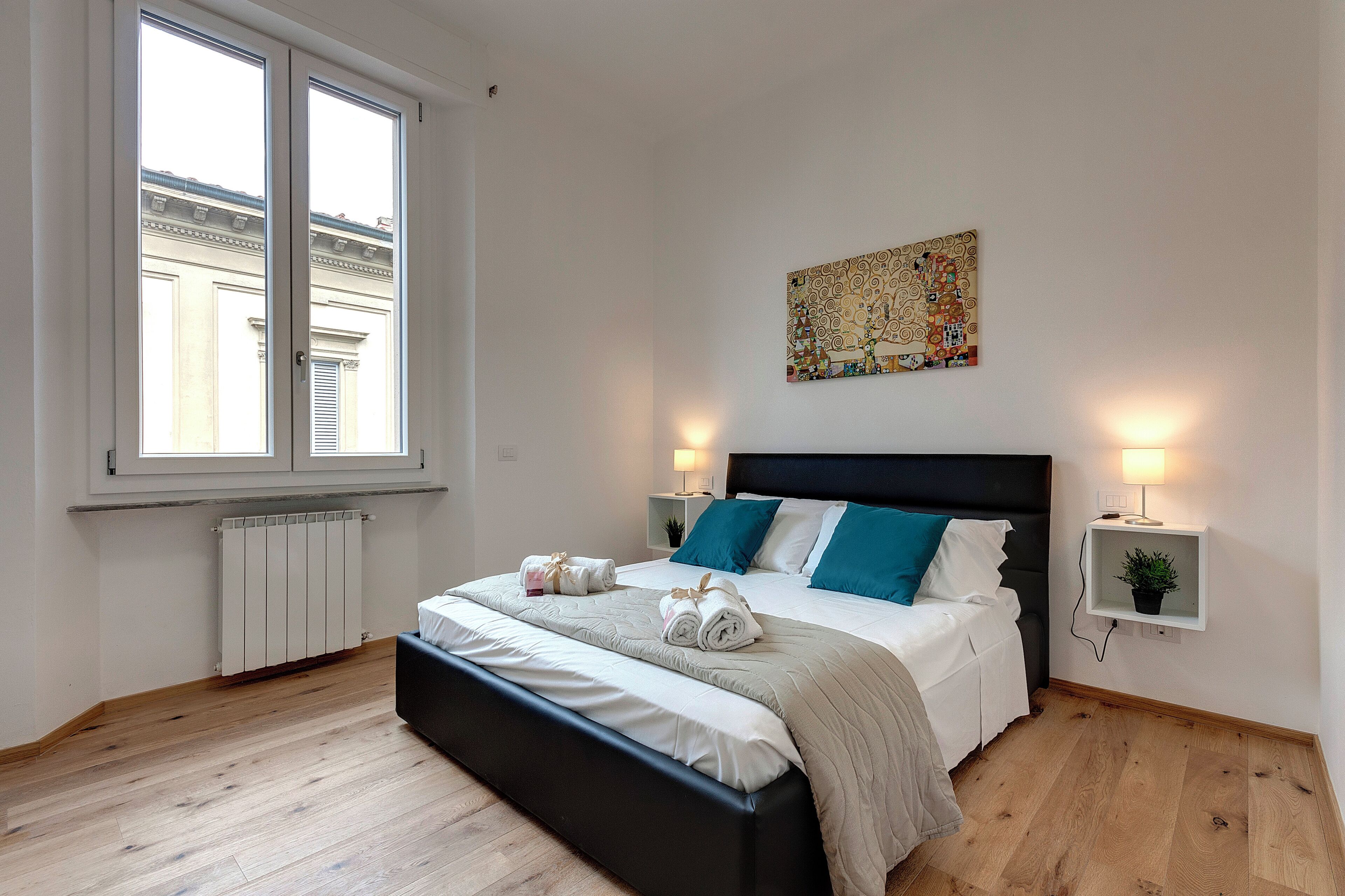 A modern bedroom with a large bed, two bedside lamps, a radiator, and a window with a view of a building.