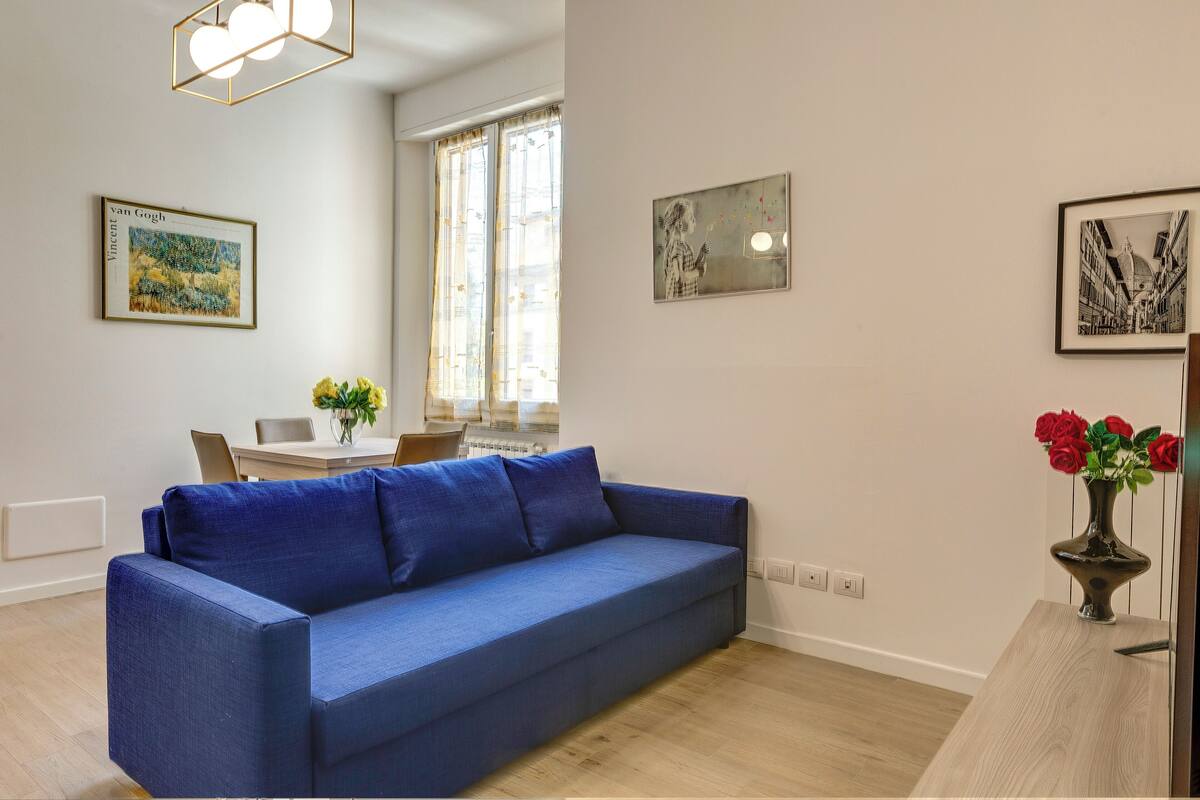 A modern living room with a blue sofa, a dining table with chairs, and artwork on the walls.