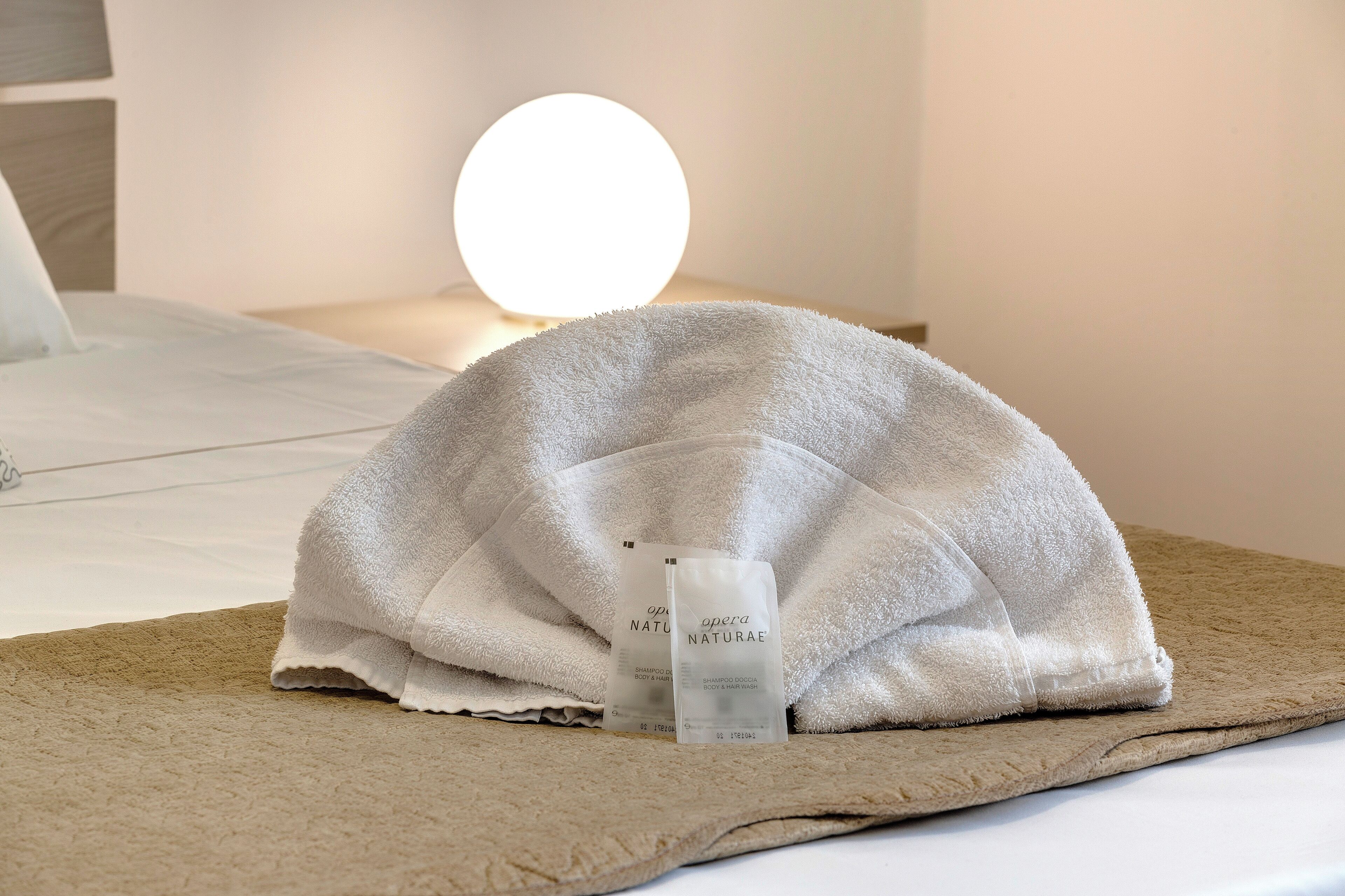 Ein ordentlich gemachtes Bett mit einem weißen Handtuch darüber, ein runder Spiegel und zwei Shampooflaschen auf dem Bett.
