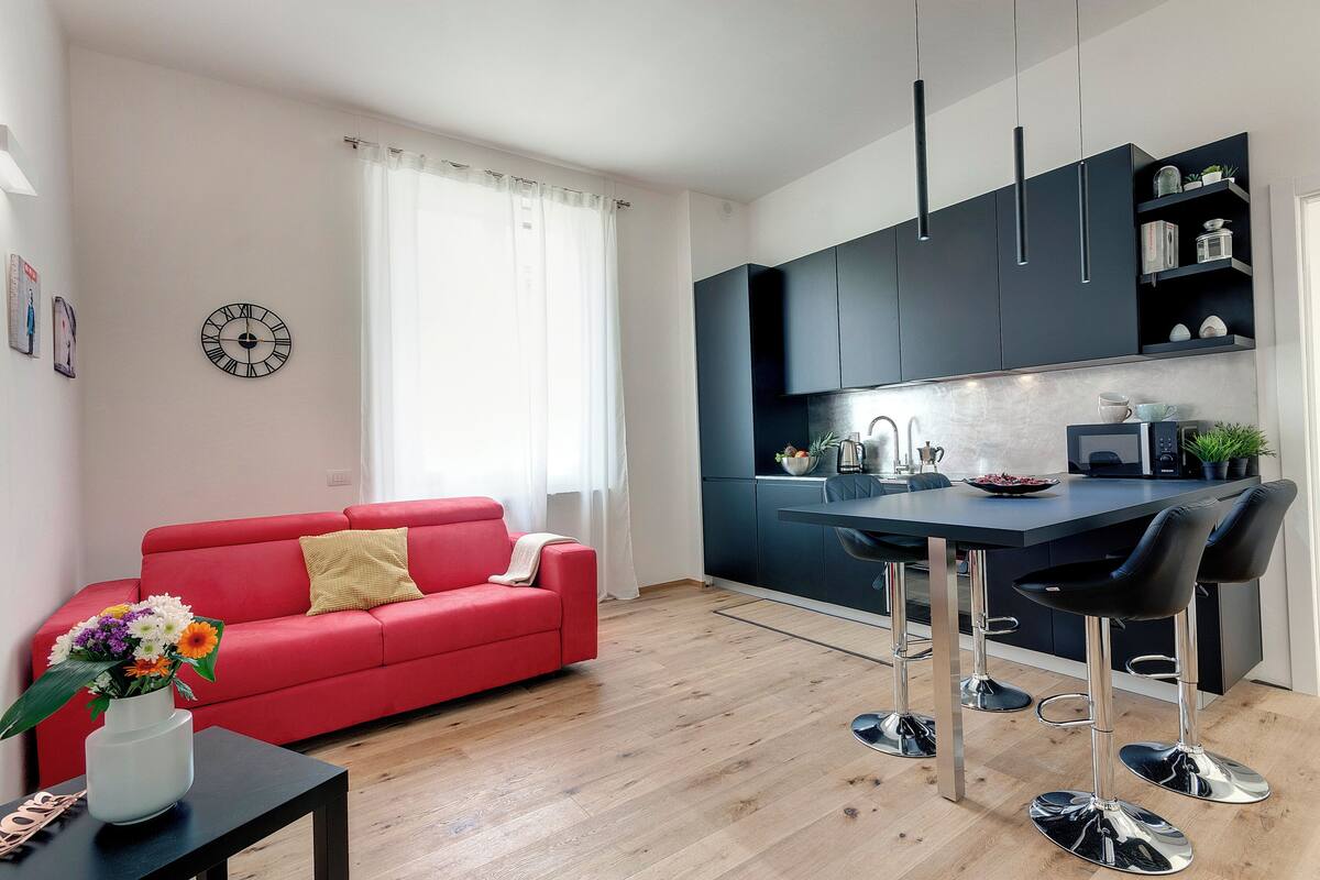 A modern living room with a red sofa, a coffee table with a vase of flowers, a kitchen with dark cabinets and a bar stool.
