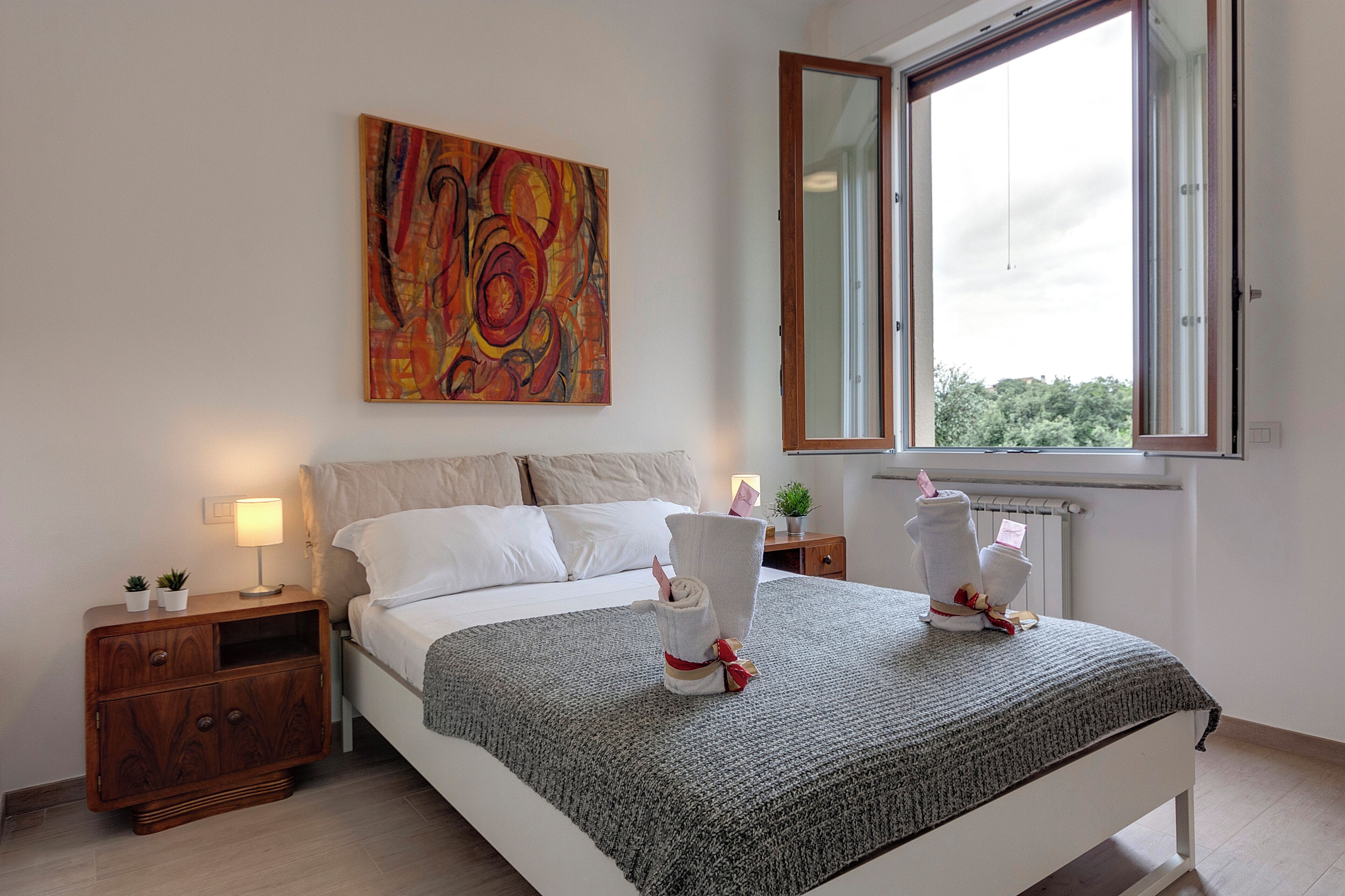 A bedroom with a bed, a nightstand, a lamp, a painting, and a window.