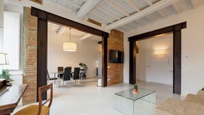 A spacious living area with a high ceiling, exposed beams, a brick accent wall, a glass coffee table, and a sofa. - Mamo Florence - Brancacci Apartment (Florence)