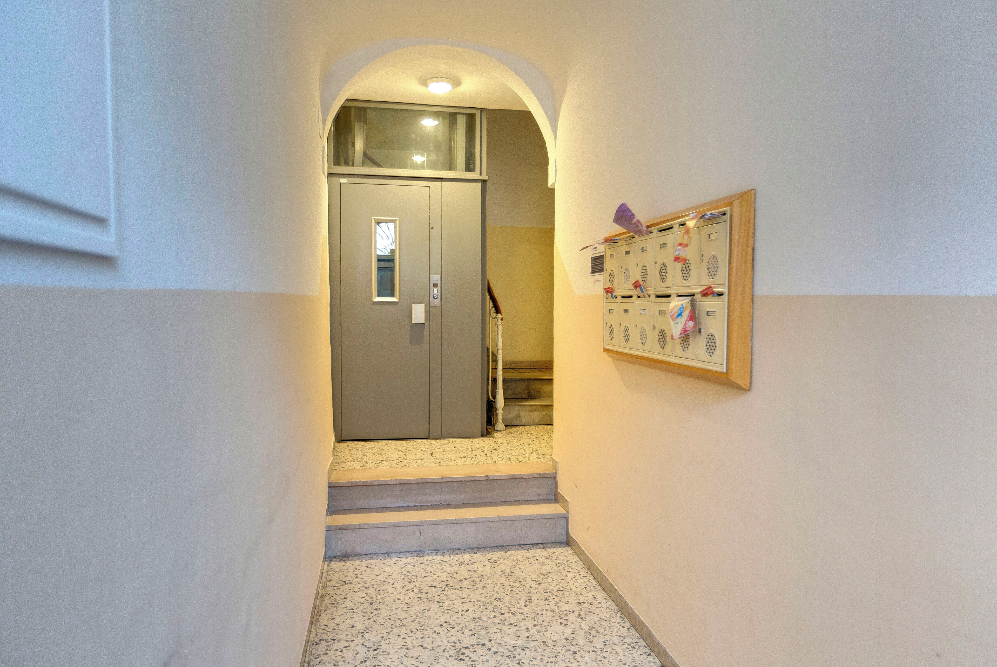 Un corridoio con una porta che dà su una scala, una bacheca con dei fogli e una lampada a soffitto.