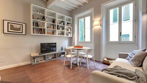 A living room with a wooden floor, a white sofa, a dining table with chairs, a bookshelf, and a TV. - Mamo Florence - Cloe Apartment (Florence)