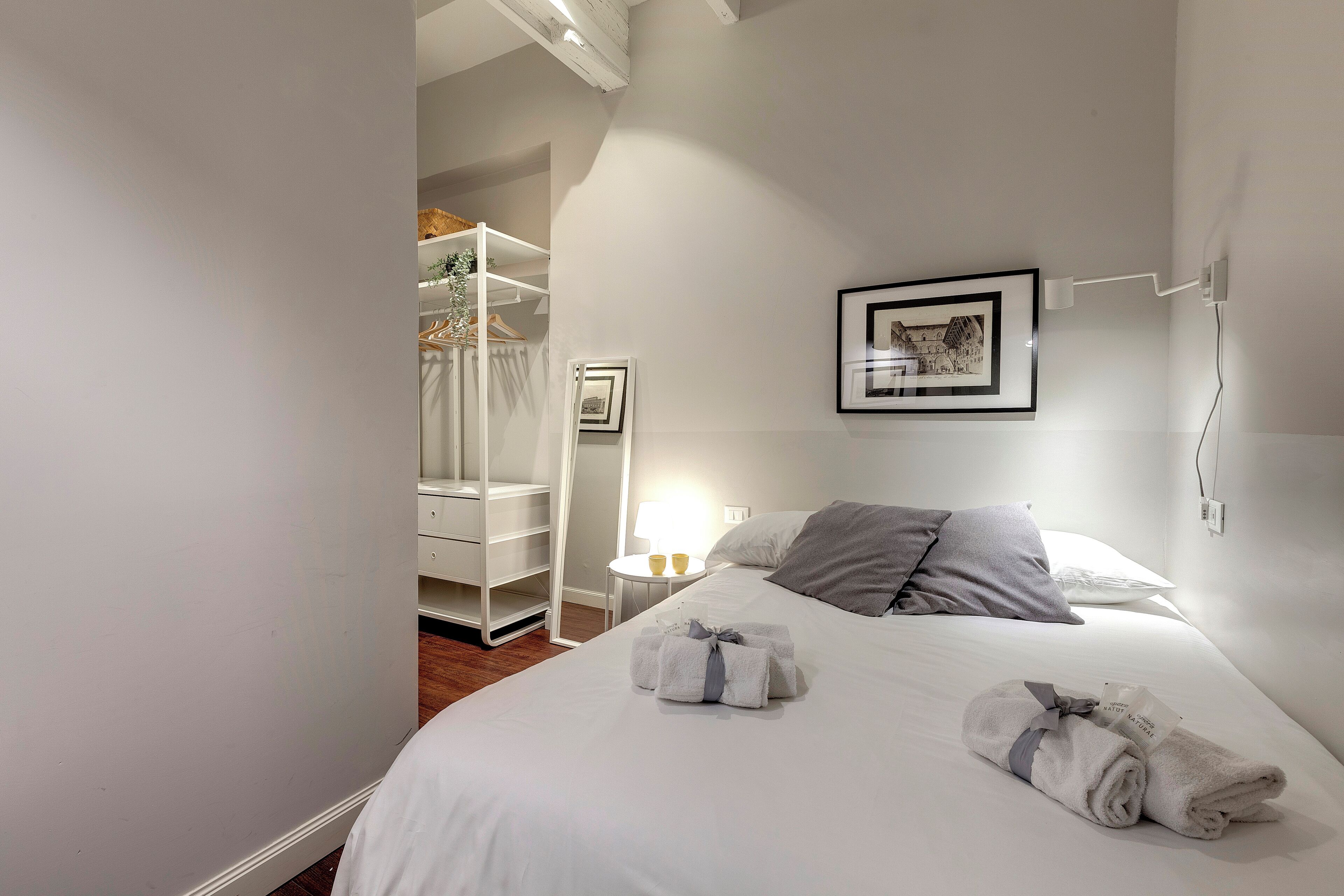 A neatly made bed with towels, a bedside table with a lamp, a framed picture on the wall, and a white wardrobe.