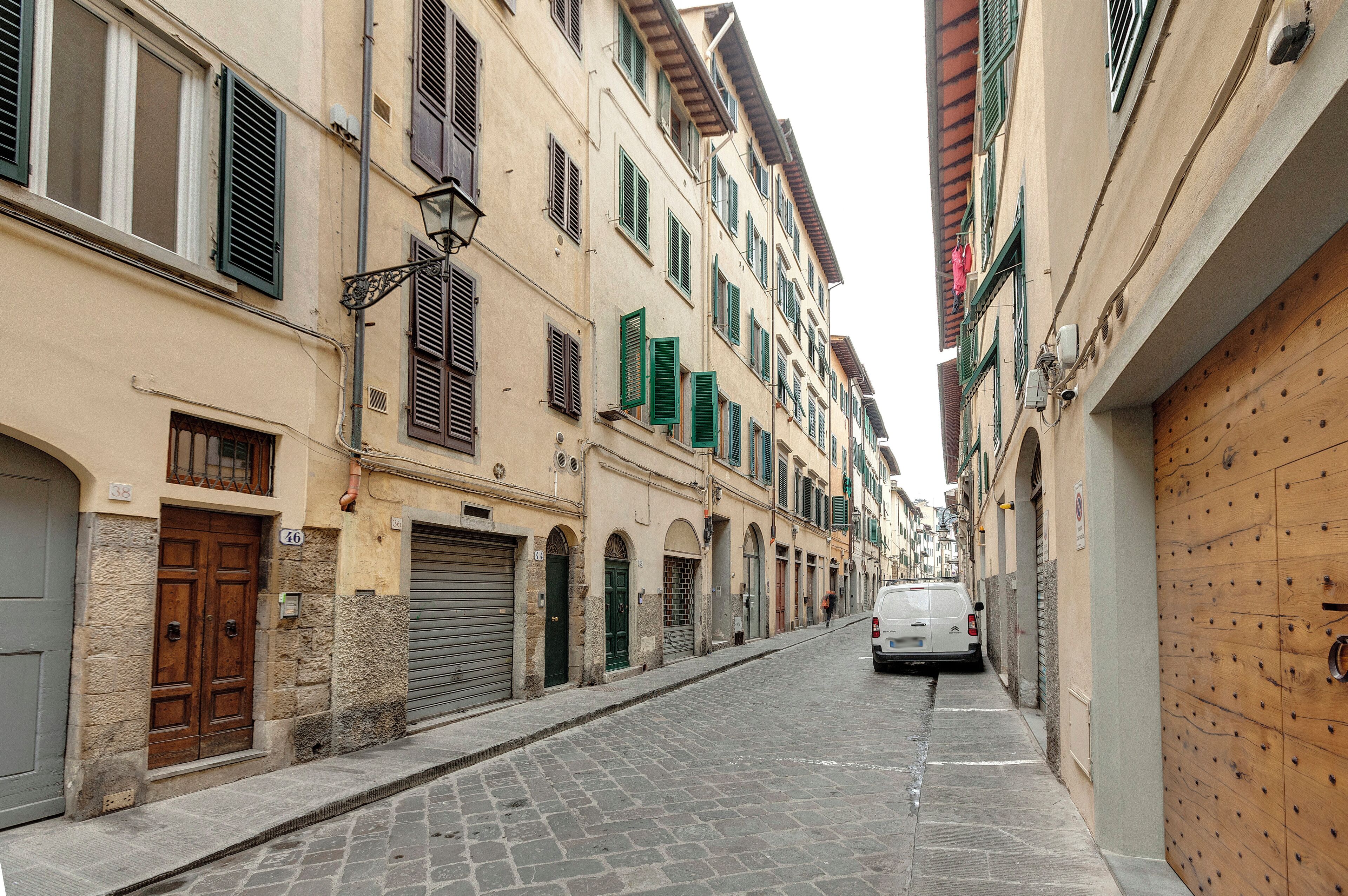 Une ruelle étroite pavée, bordée de bâtiments historiques aux façades en pierre, aux volets verts, et où une voiture blanche est garée sur le côté droit.