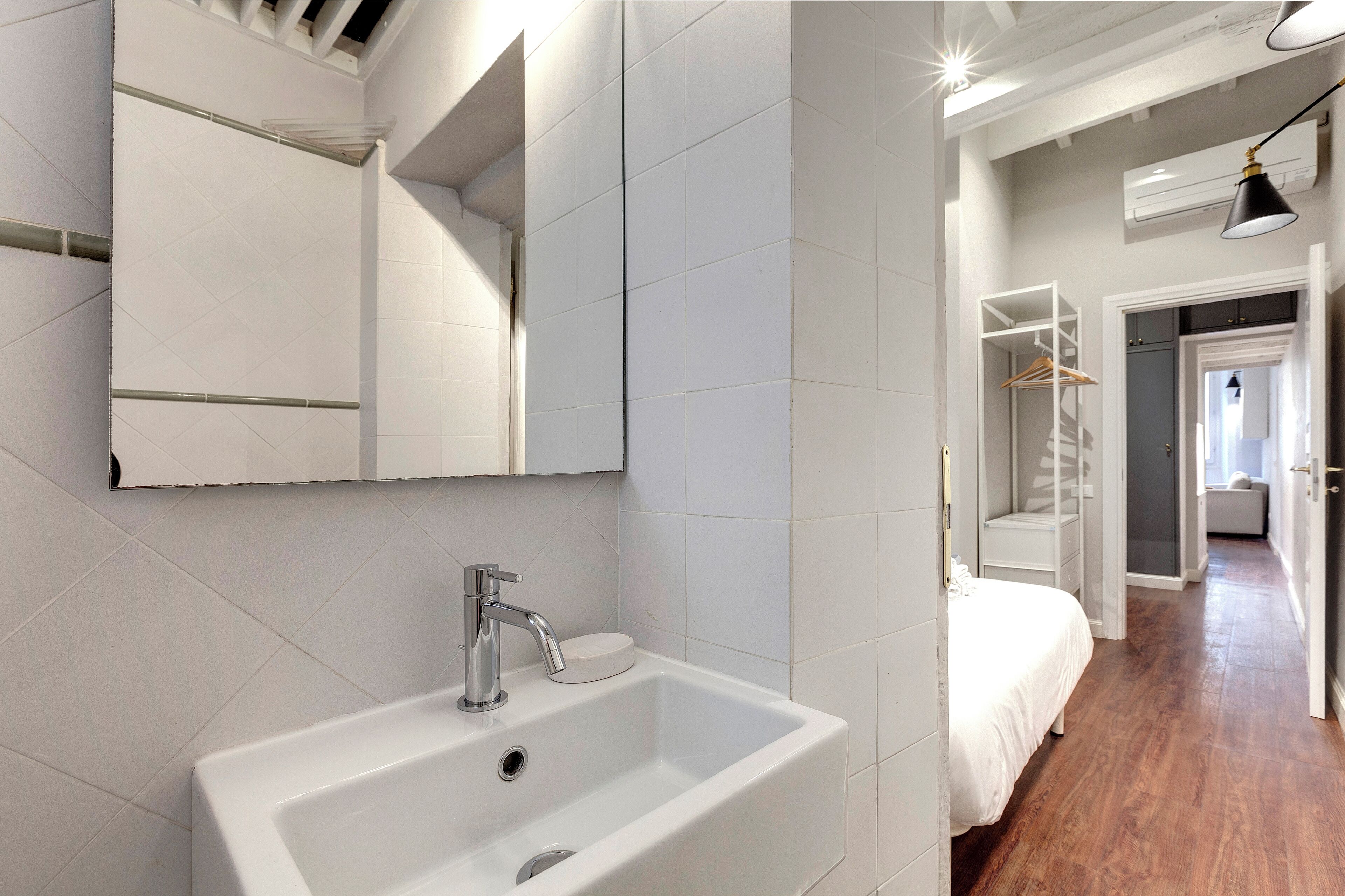 Une salle de bain moderne avec un lavabo blanc, un miroir et des murs carrelés. Le sol est en bois et un couloir mène à une chambre à coucher équipée d’un lit et d’un placard.