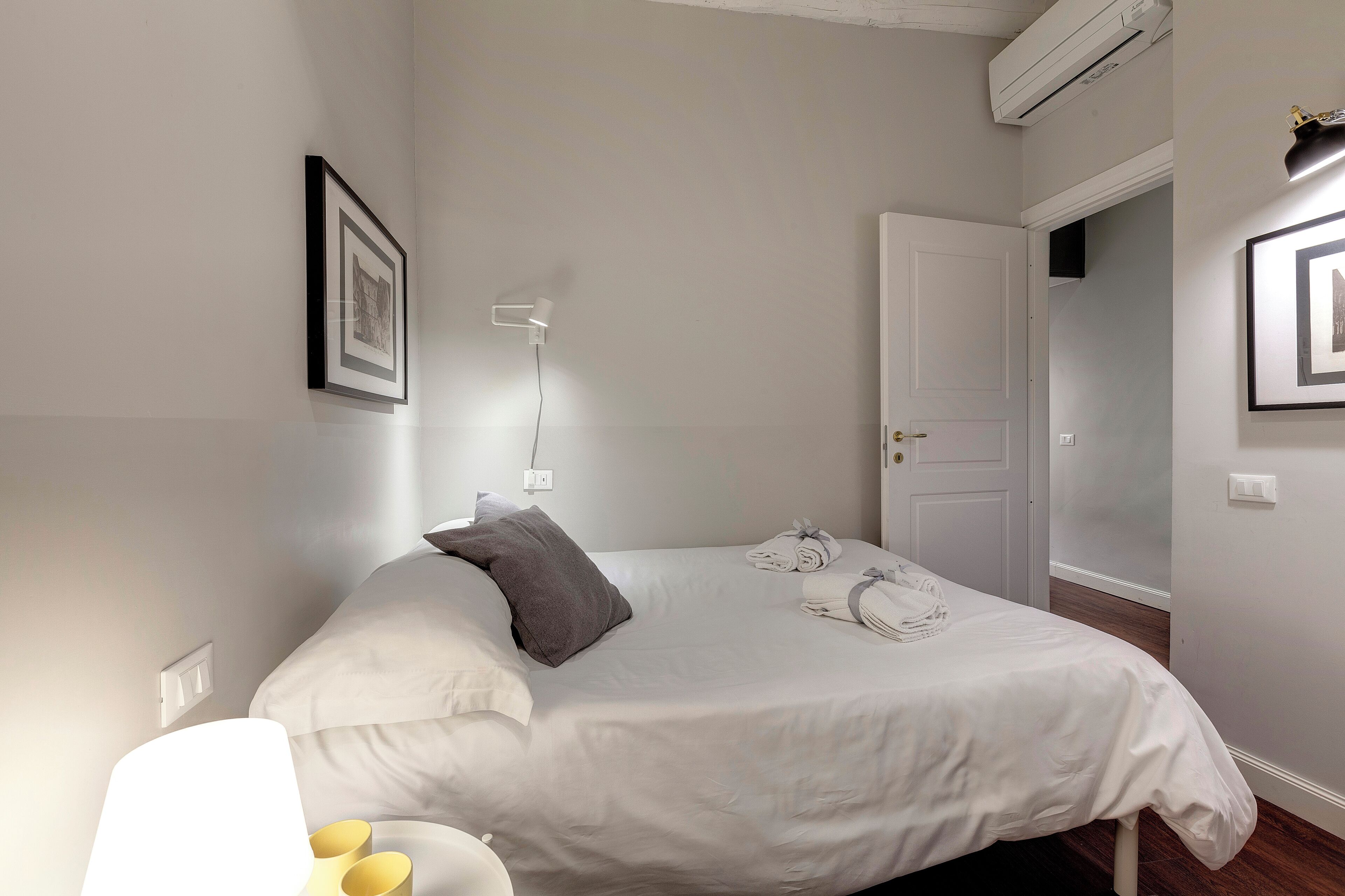 A neatly made bed with white bedding and a gray pillow, a bedside lamp, and a framed picture on the wall.