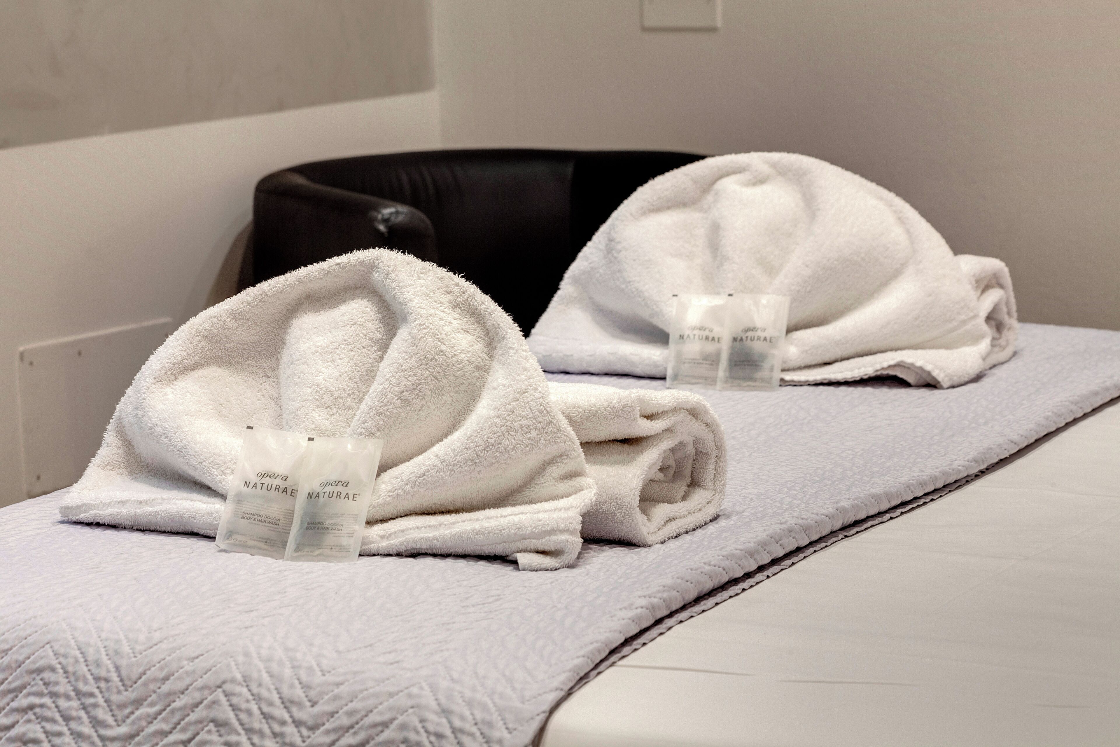 Ett hotellrum med en säng täckt av ett vitt handduk och två flaskor av märket ”Opera Natura” på sängen.