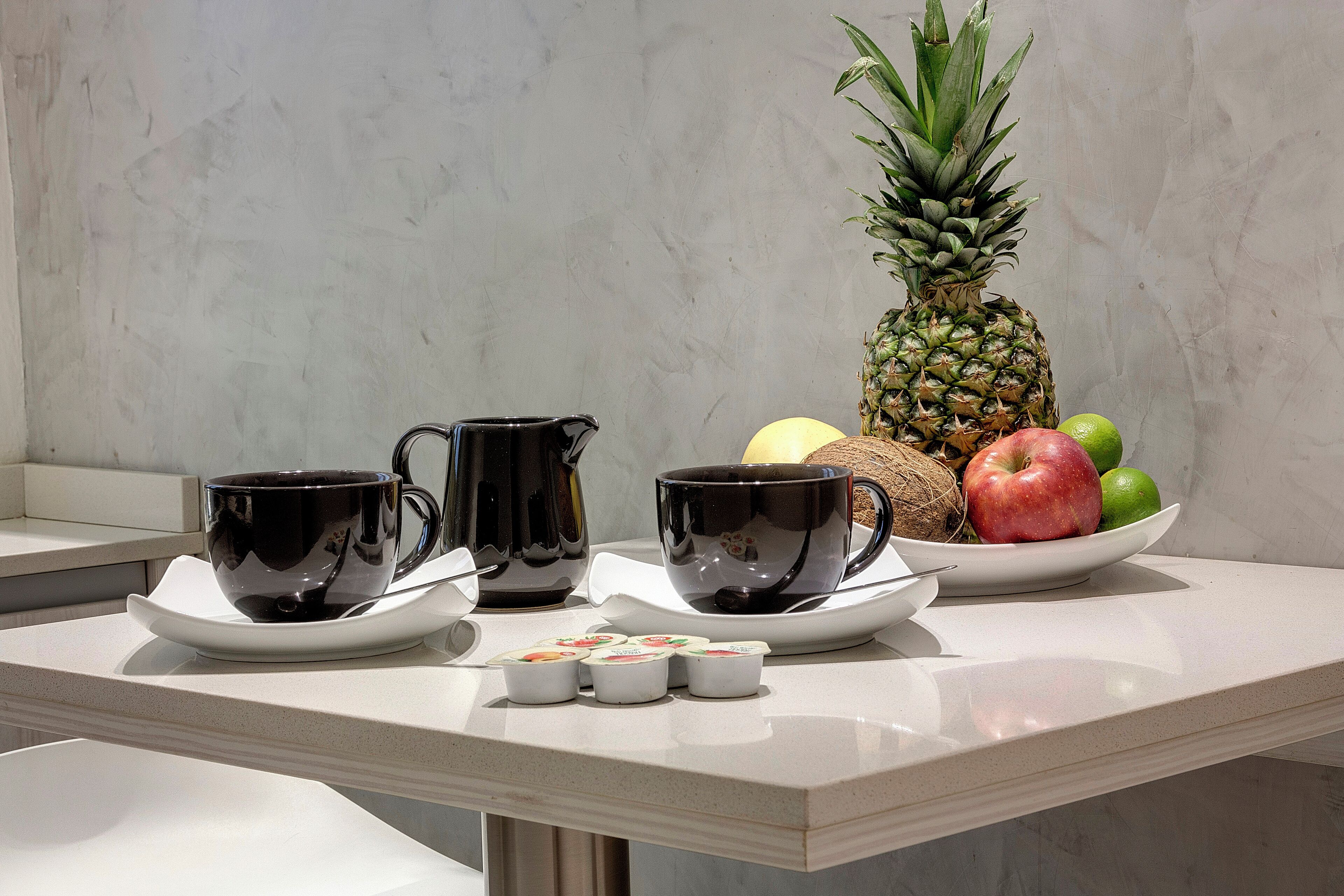 A kitchen counter with two black coffee cups on saucers, a pineapple, and a bowl of fruit including apples and limes.