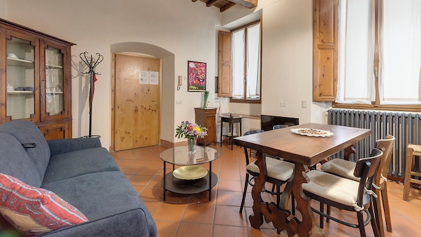 A cozy living room with a sofa, a dining table, and chairs. There is a wooden cabinet with glass doors, a vase with flowers, and a radiator. - Mamo Florence - Dafne Apartment (Florence)