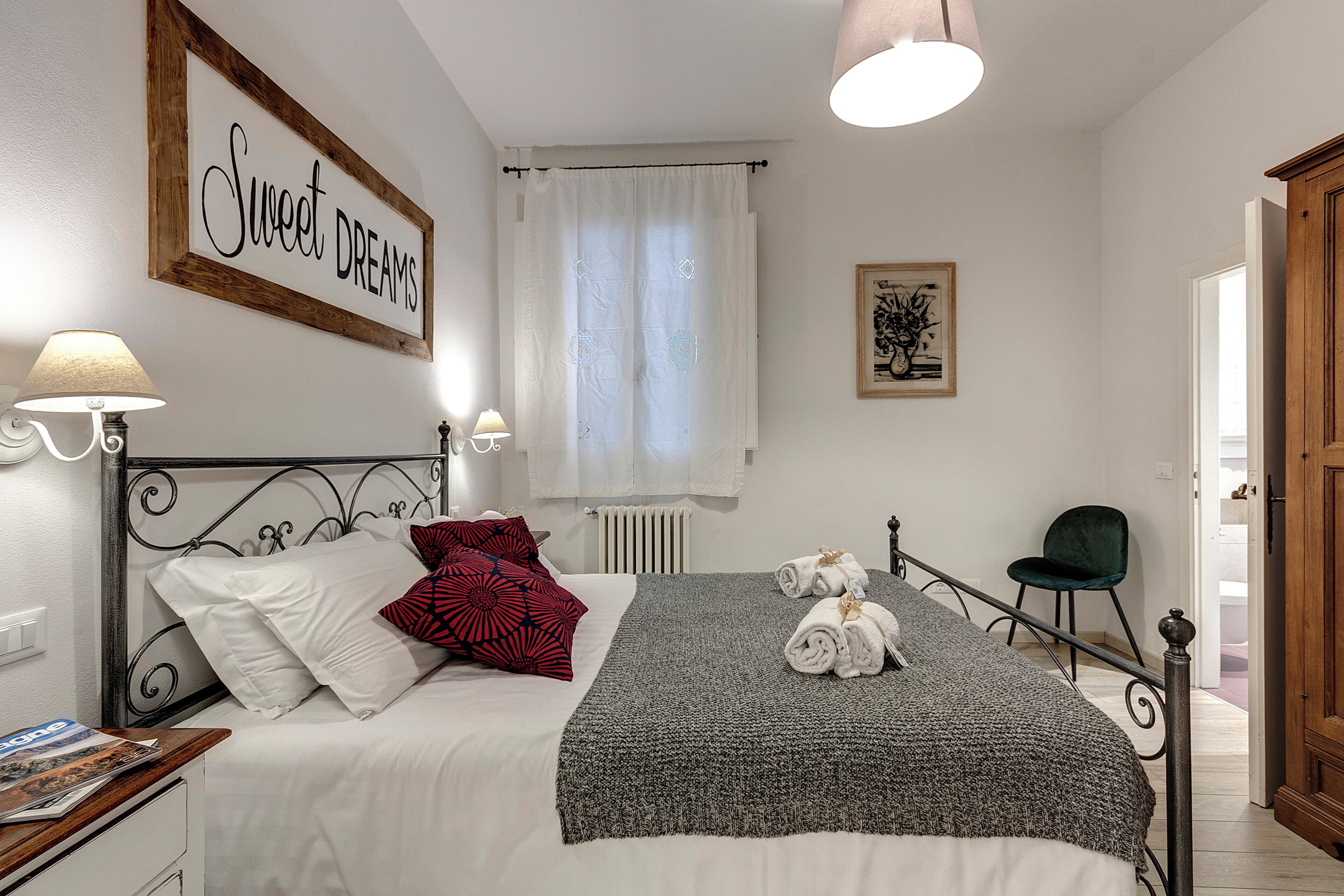 A bedroom with a bed, pillows, a bedside table, a lamp, a window with curtains, a framed artwork, and a chair.