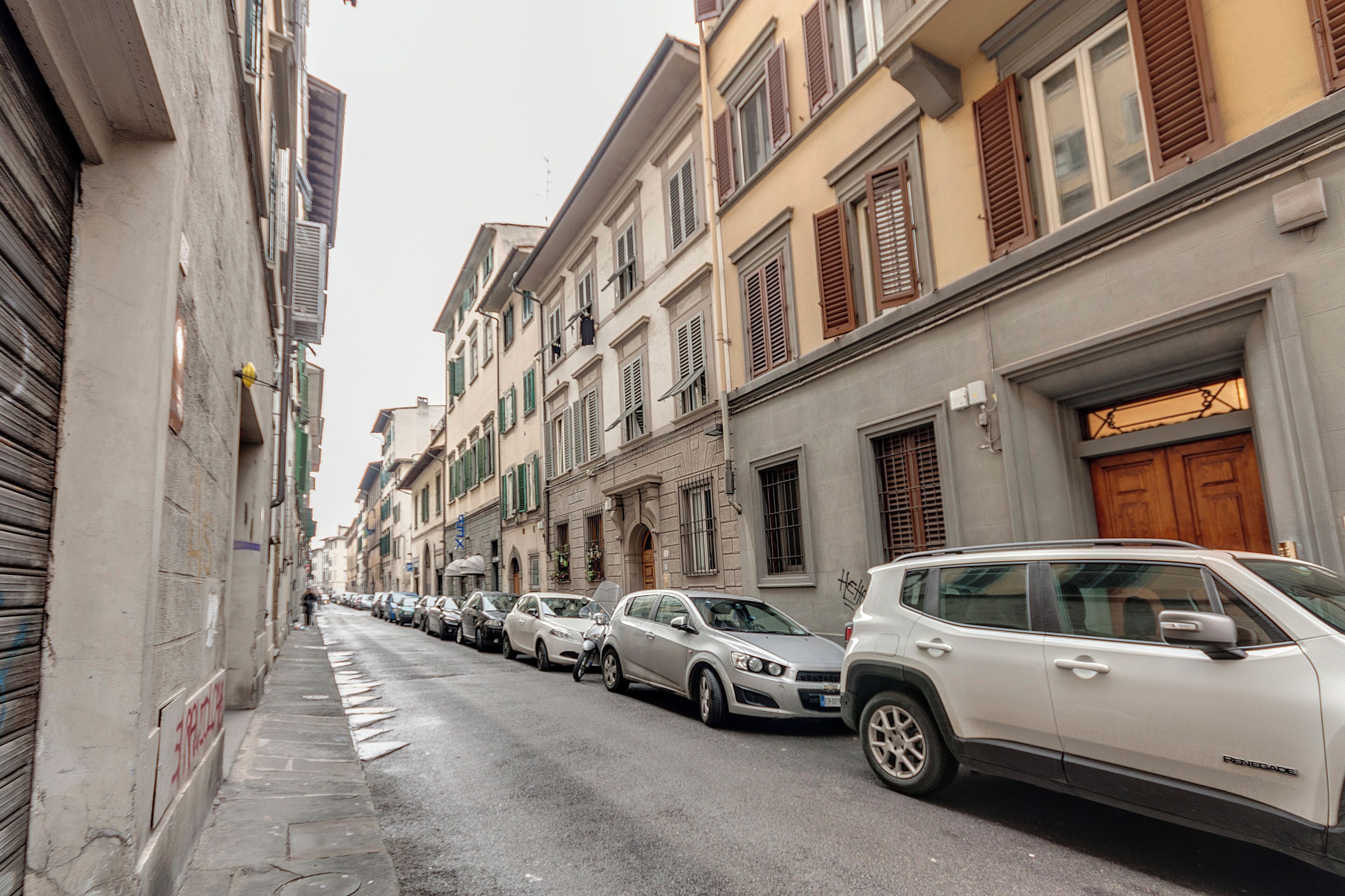 Une rue étroite bordée de voitures garées et d’immeubles aux volets fermés.