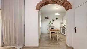 Un couloir avec une arche en briques menant à une salle à manger équipée d’une table et de chaises en bois, d’un luminaire suspendu et offrant une vue sur une cuisine avec des placards et des appareils électroménagers.