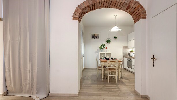 Un couloir avec une arche en briques menant à une salle à manger équipée d’une table et de chaises en bois, d’un luminaire suspendu et offrant une vue sur une cuisine avec des placards et des appareils électroménagers.