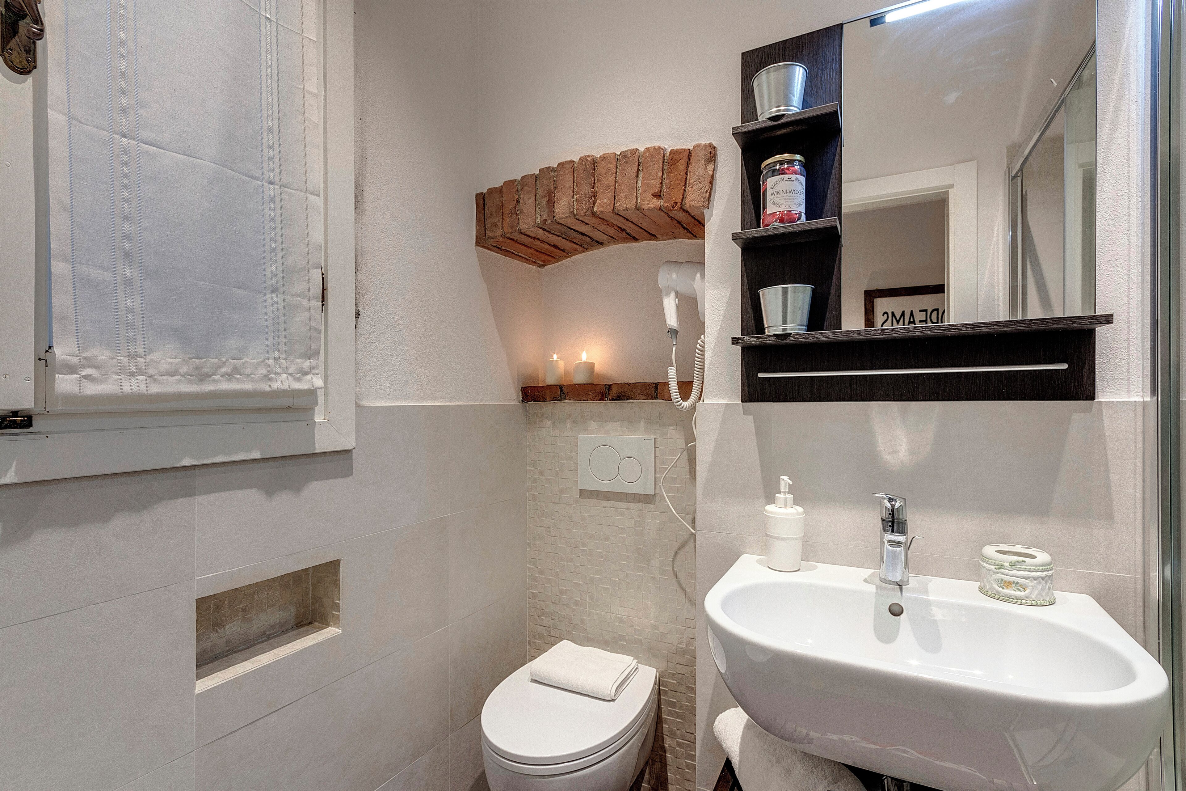 A bathroom with a toilet, sink, and a mirror above the sink. There is a shelf with a jar and a candle on it.