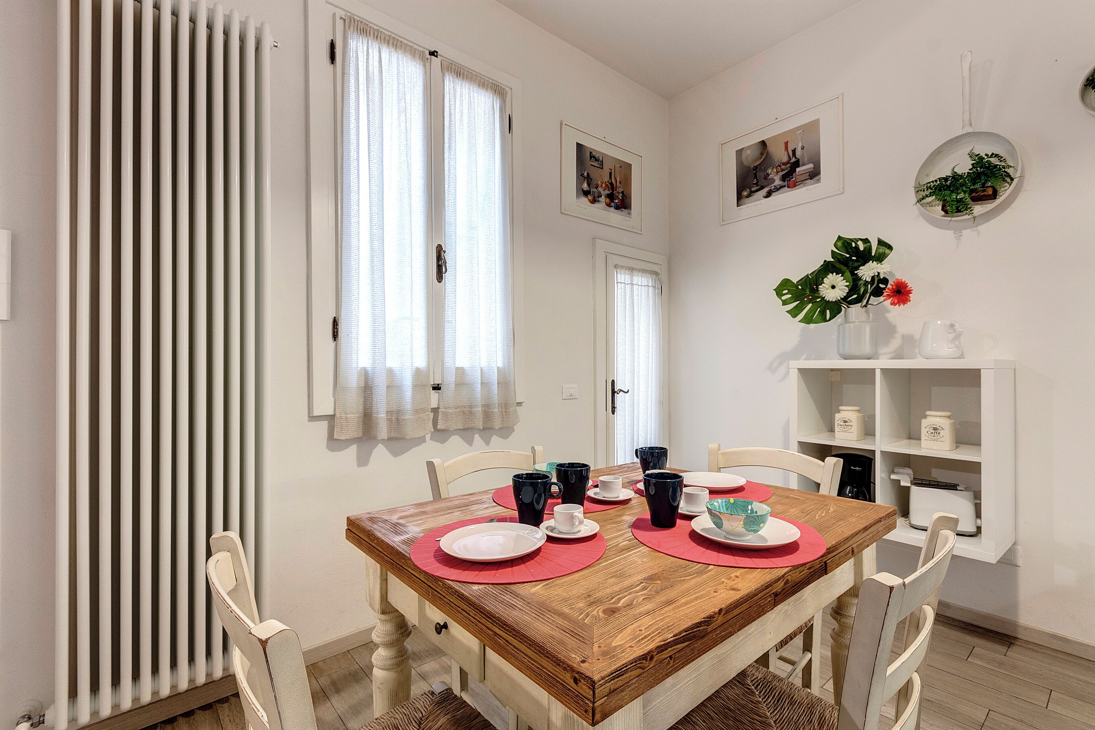 A dining area with a wooden table set for four, white chairs, a radiator, and a shelf with kitchen items.