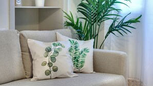 Un canapé beige agrémenté de deux coussins décoratifs ornés de motifs de feuillage vert. Une grande plante verte est placée à droite du canapé, et une étagère blanche avec une plante en pot est visible à l’arrière-plan.