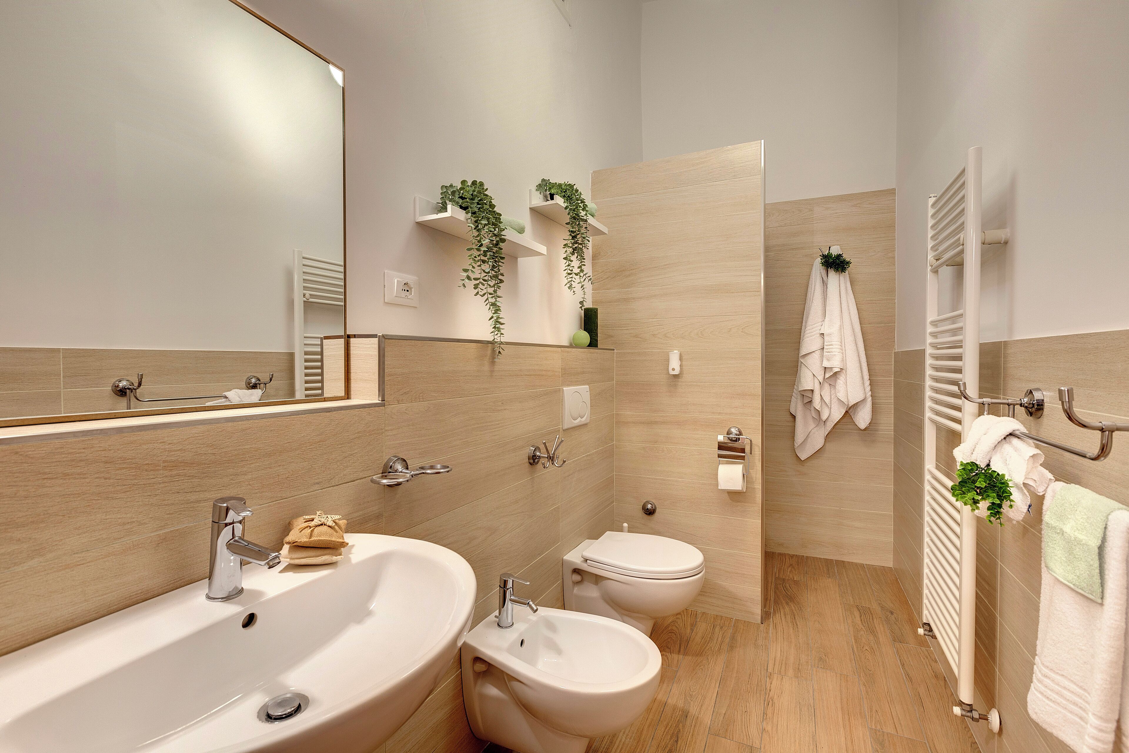 Ein modernes Badezimmer mit einem großen Waschbecken, einem Bidet und einer Toilette. Pflanzen stehen auf Regalen und ein Handtuchhalter mit Handtüchern ist vorhanden.