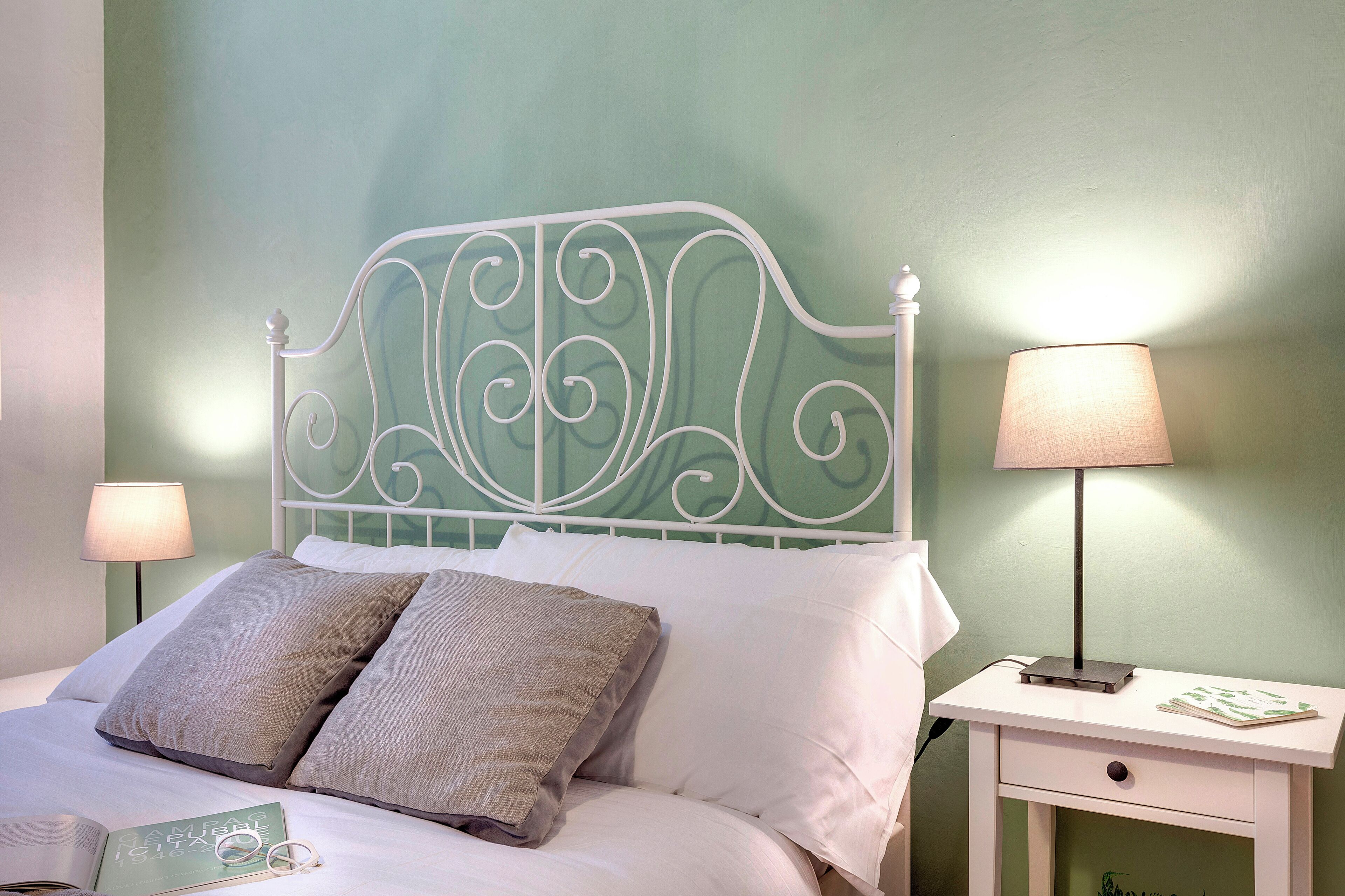 A neatly made bed with white linens and two pillows, a white metal headboard, a bedside table with a lamp, and a small drawer.