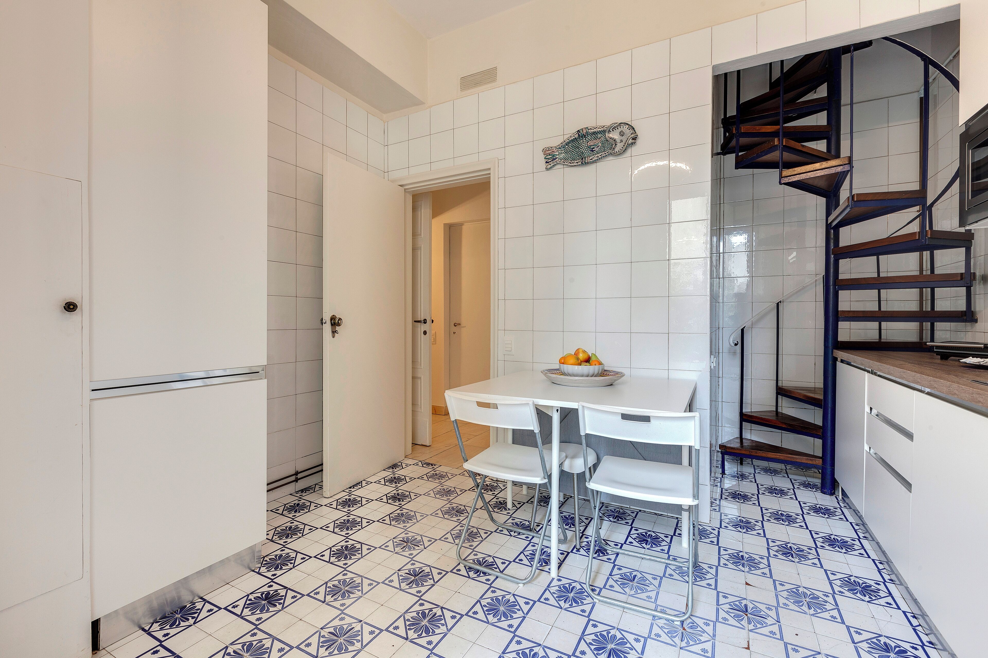 A kitchen with white cabinets, a white table, blue and white patterned floor tiles, and a spiral staircase.