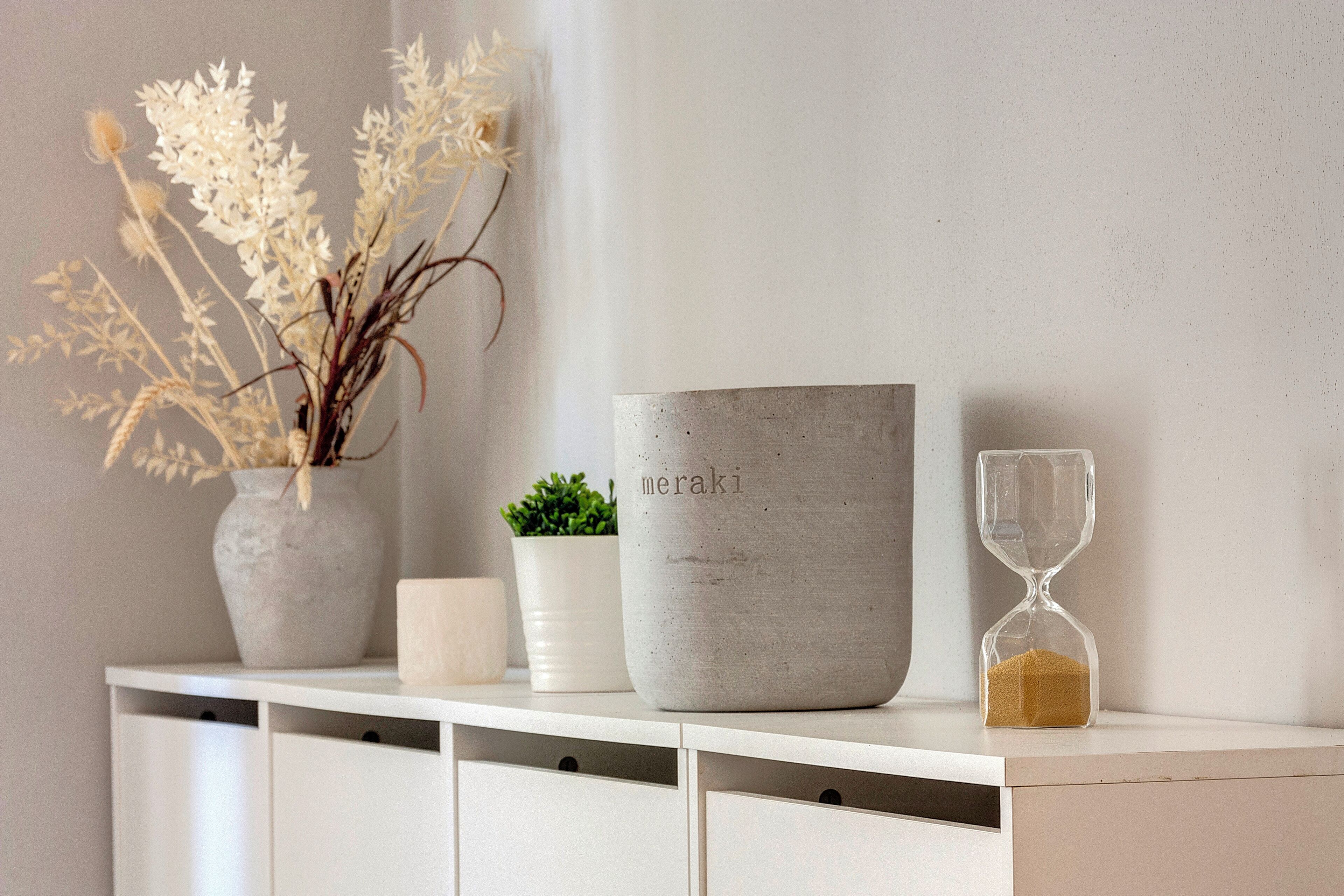 A white console table with a vase of white flowers, a potted plant, a large grey container labeled "meraki," and an hourglass.