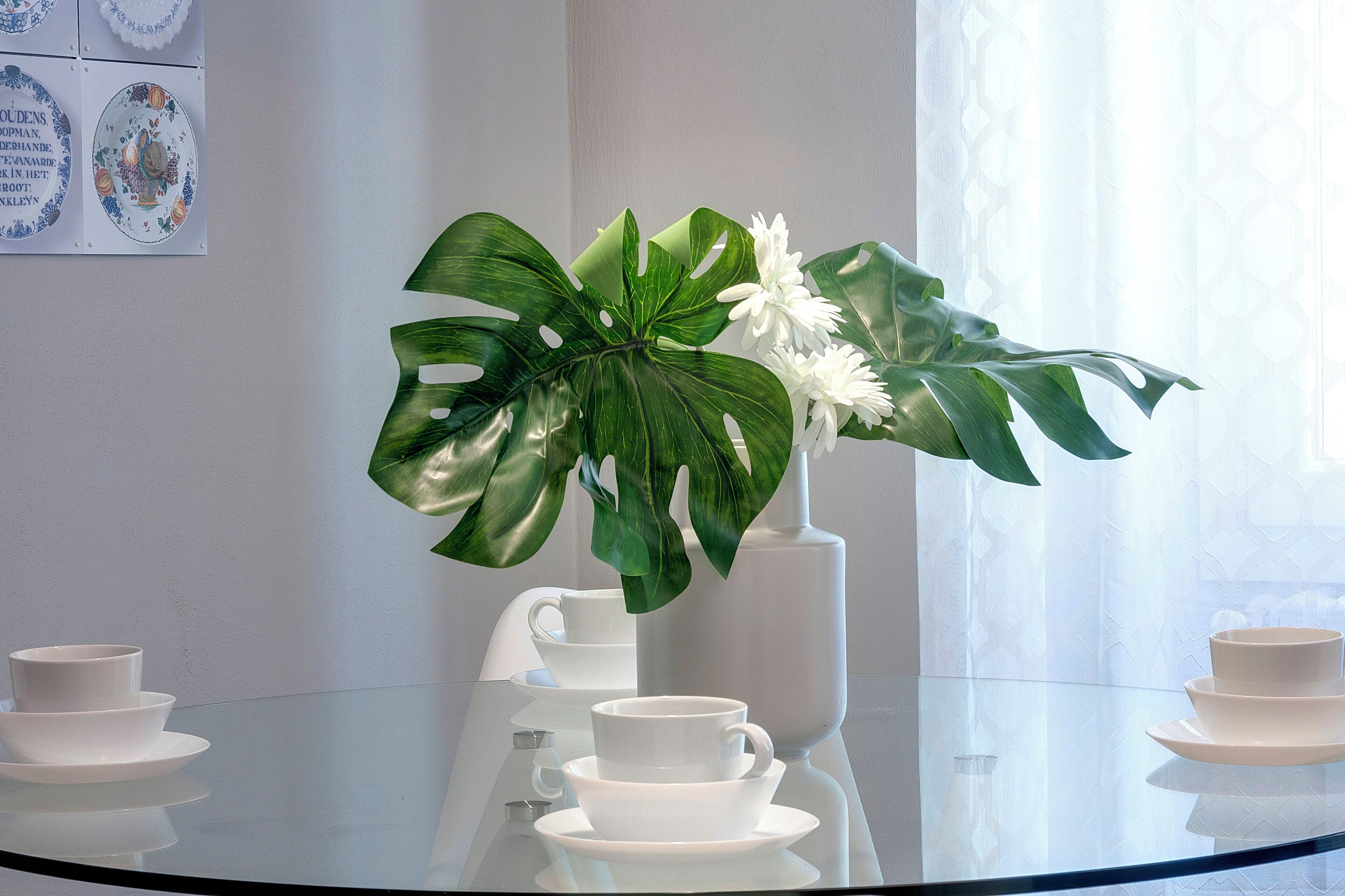 Een glazen tafel met witte kopjes en schotels, een witte vaas met witte bloemen, en een grote groene plant.