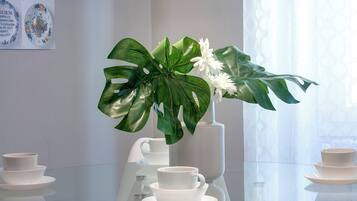 Een glazen tafel met witte kopjes en schotels, een witte vaas met witte bloemen, en een grote groene plant.