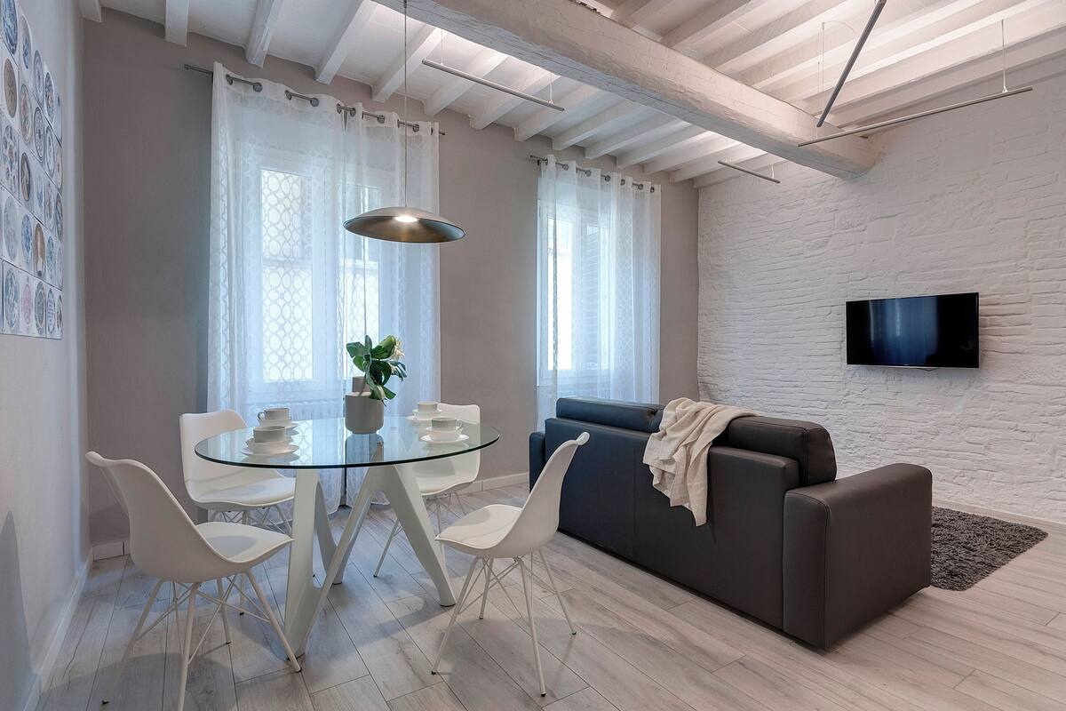 Un salon moderne avec une table à manger en verre, des chaises blanches, un canapé foncé et un téléviseur à écran plat fixé à un mur en briques blanches.