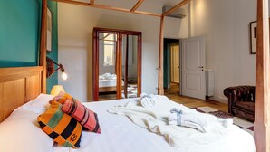 A bedroom with a wooden headboard, a bed with white bedding, a colorful pillow, a white blanket, and a brown leather chair. - Mamo Florence - Frida Luxury Apartment (Florence)