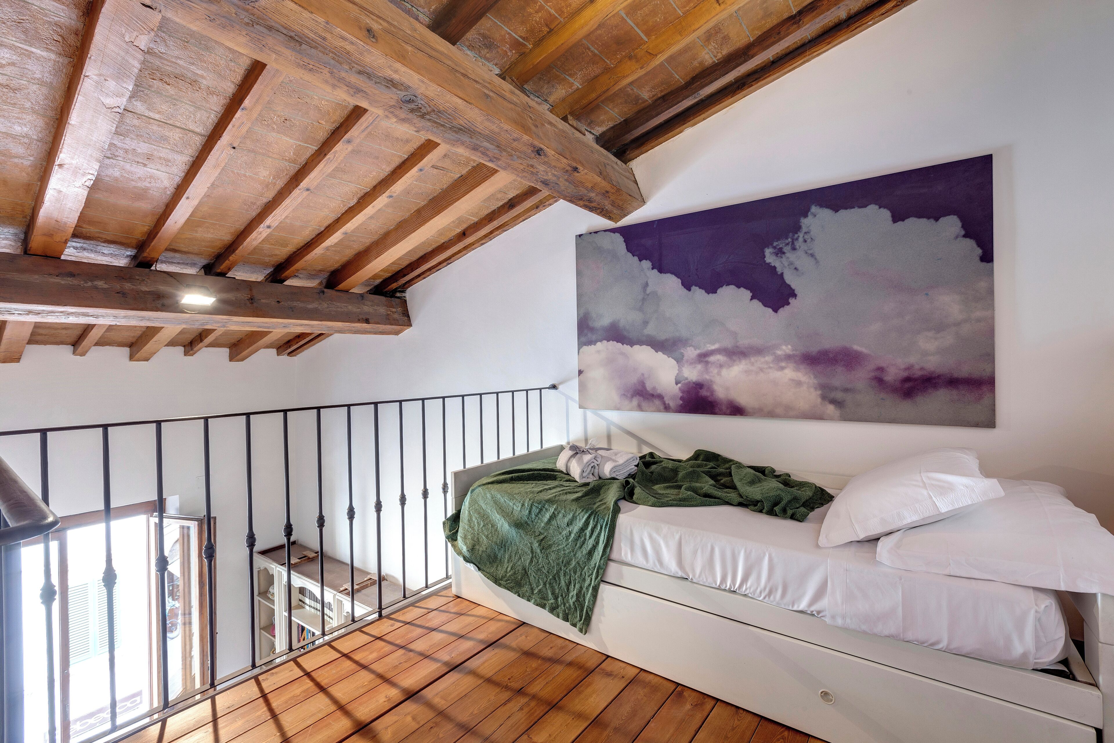 A loft bedroom with exposed wooden beams, a white bed, a green blanket, and a purple cloud painting on the wall.