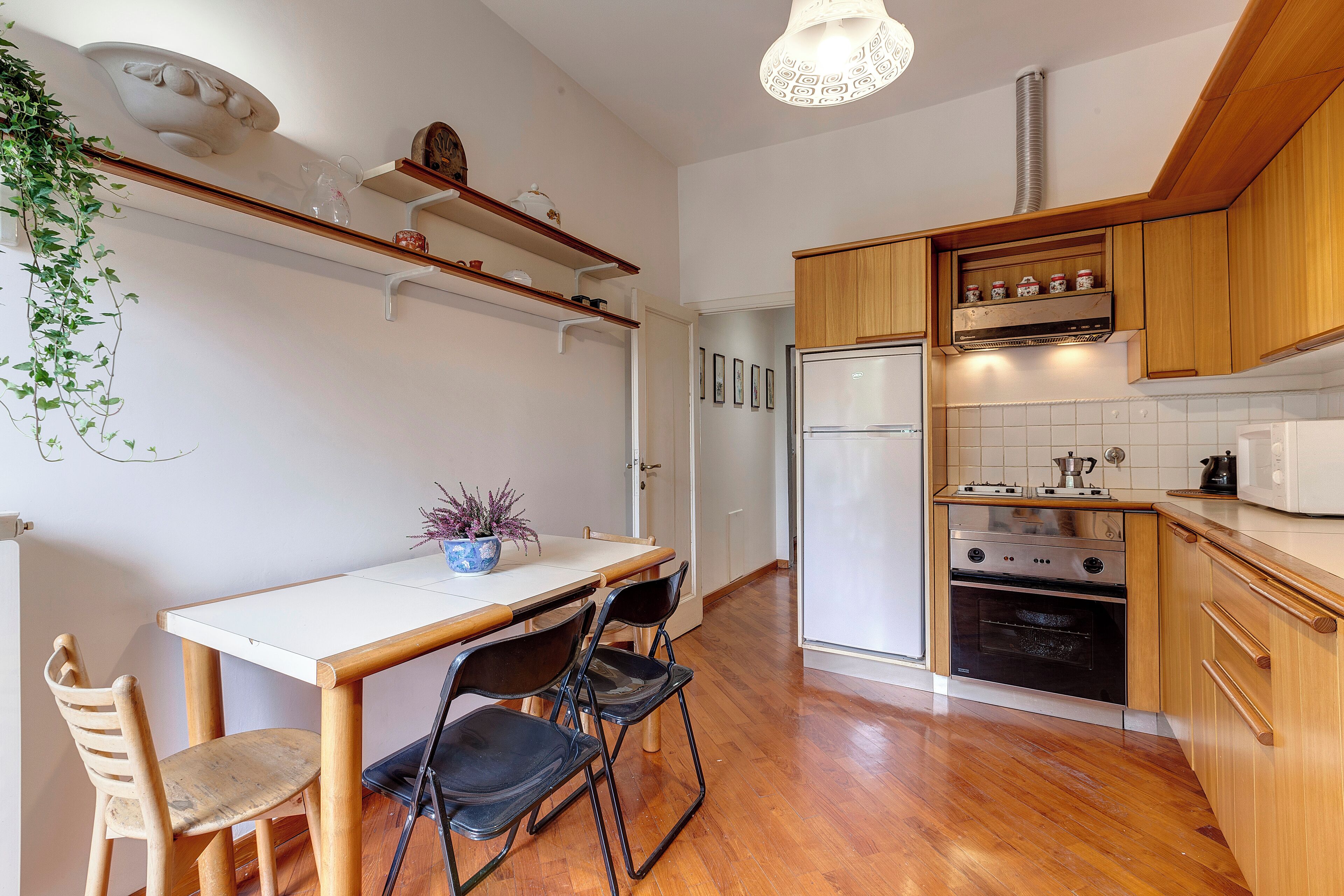 A compact kitchen with wooden cabinets, a built-in oven, and a refrigerator. There is a dining table with chairs and a potted plant on the table.