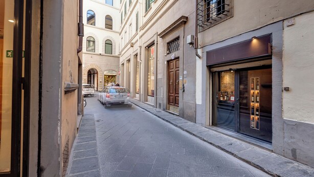 A narrow street with buildings on either side, a car parked on the left, and a storefront with illuminated bottles. - Mamo Florence - Guendalina Apartment (Florence)
