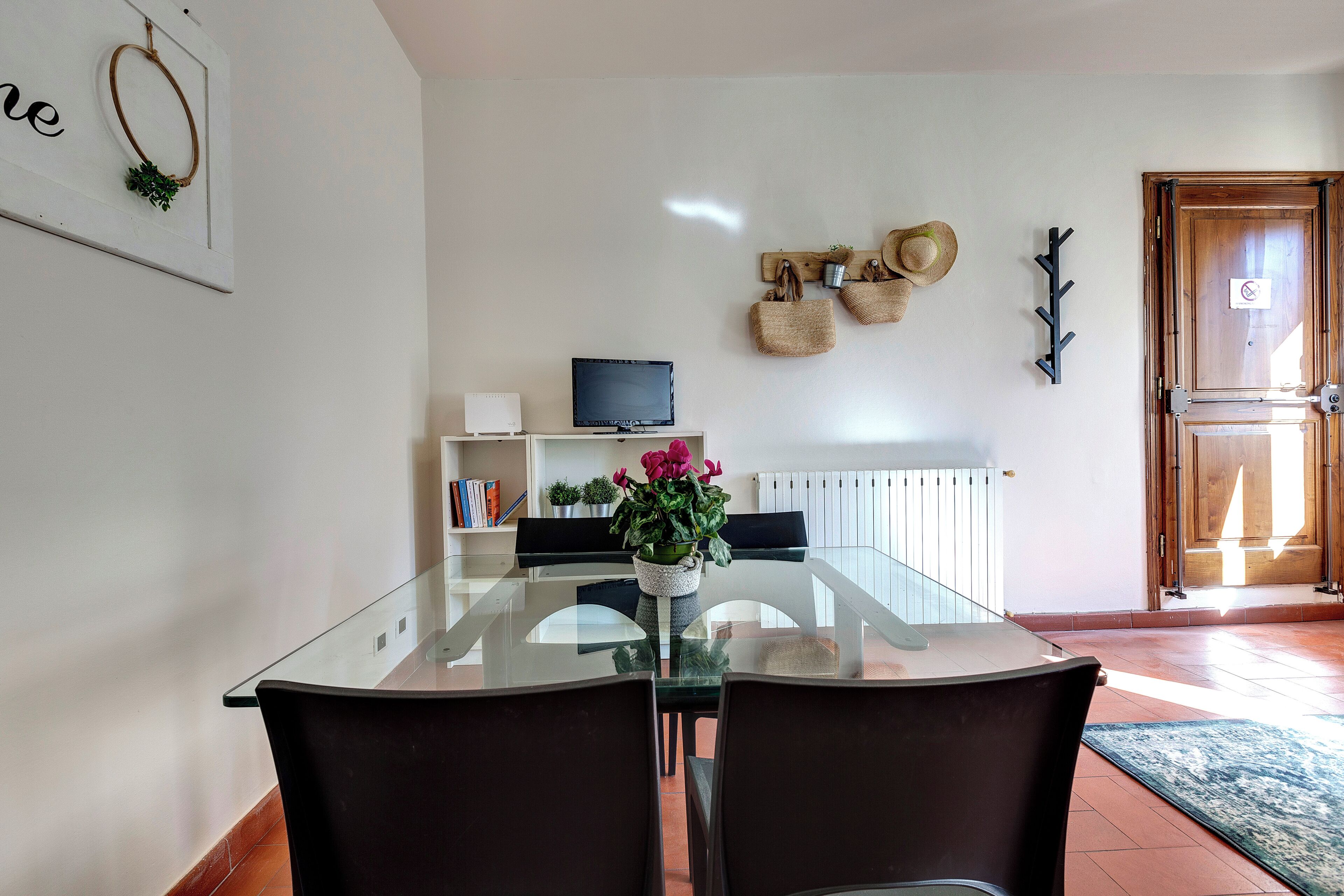 A modern dining area with a glass table and dark chairs, a television on a shelf, and a door with a mat.