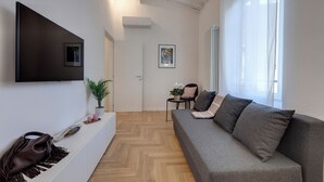 Een moderne woonkamer met een grijze bank, een wandtelevisie, een kleine tafel met een plant en een witte console met een handtas en kaarsen.