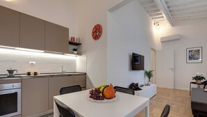 Een moderne keuken met een eetgedeelte, een witte tafel met een fruitstuk en een aan de muur bevestigde televisie.