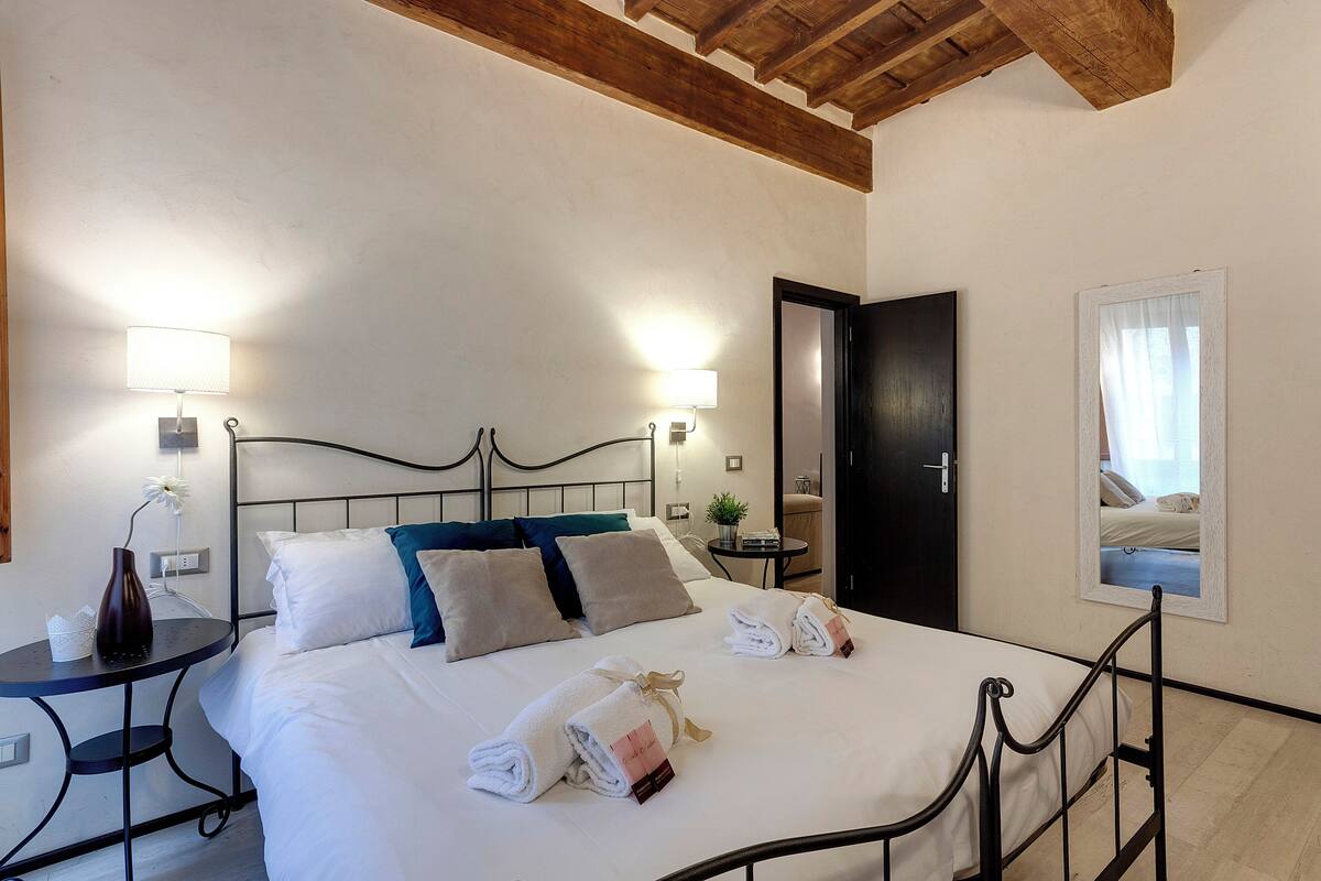 Una cama bien hecha con ropa de cama blanca y azul, dos mesitas de noche con lámparas, y un techo de madera.