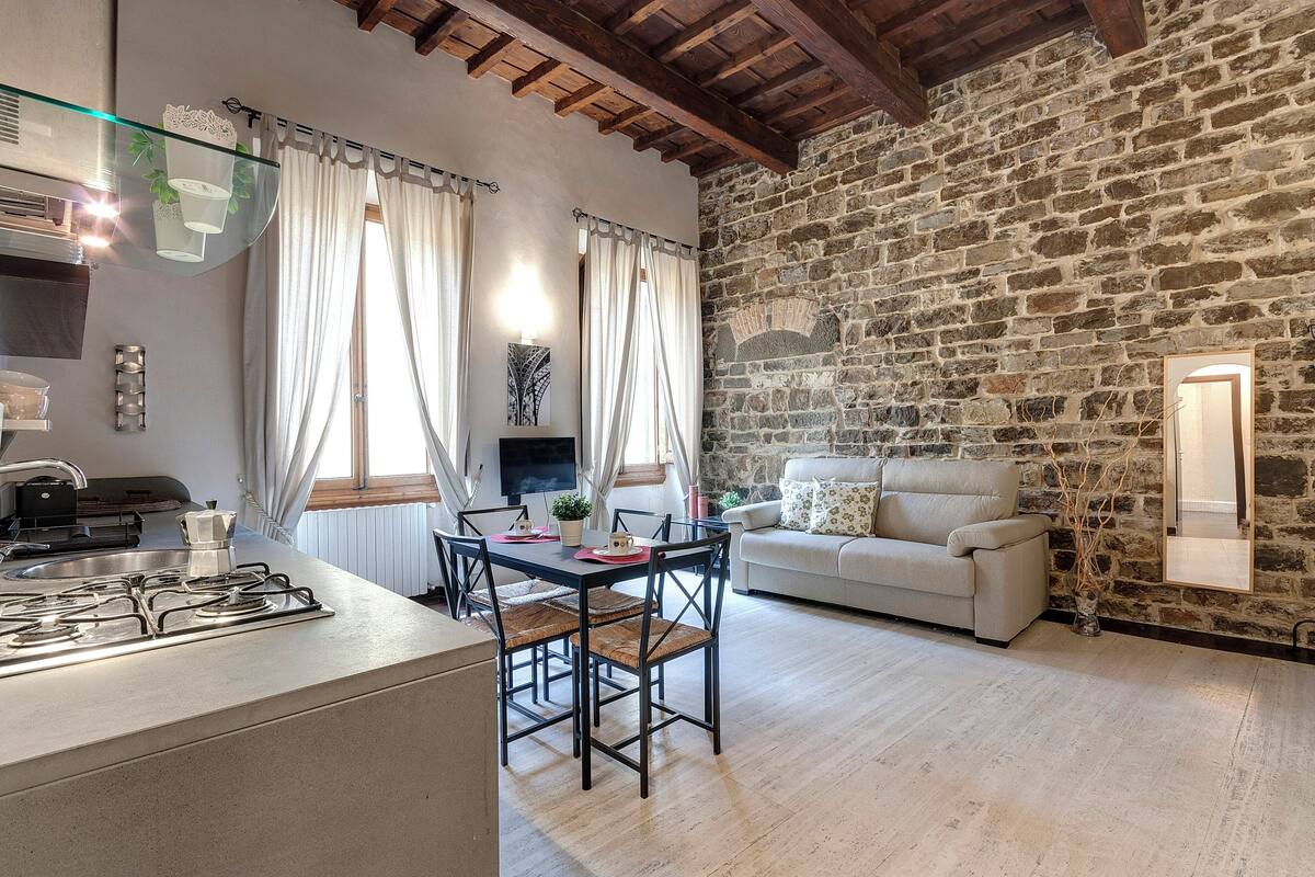 Cocina con estufa a gas, mesa de comedor y sillas. En la sala de estar, hay una pared de piedra y un sofá blanco.