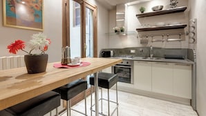 Cozinha moderna com mesa de madeira, dois bancos altos pretos, micro-ondas, forno e pia. Há itens decorativos nas prateleiras e um quadro na parede.
