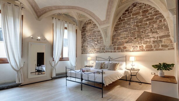 Quarto com parede de pedra, cama com estrutura de metal, mesa de cabeceira com abajur e planta em vaso.