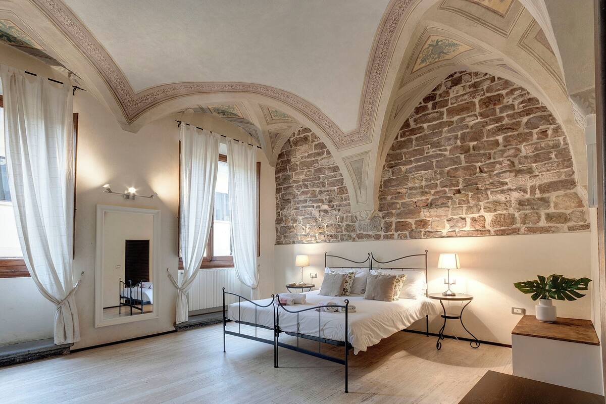 Un dormitorio con pared de piedra, una cama con estructura metálica, una mesita de noche con lámpara y una planta en maceta.