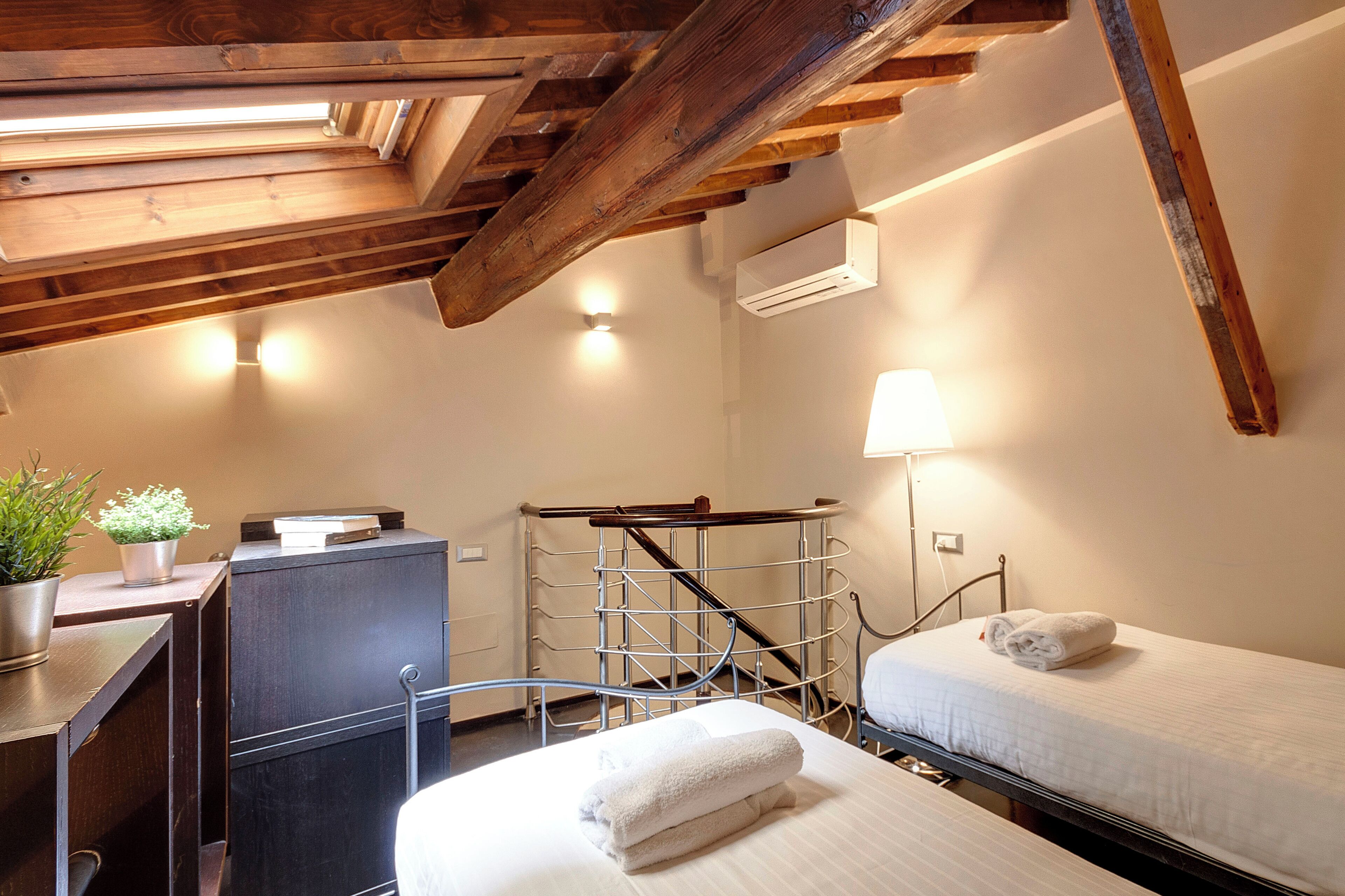 A hotel room with a wooden ceiling, a desk with a potted plant, a bed with a rolled towel, and a metal ladder.